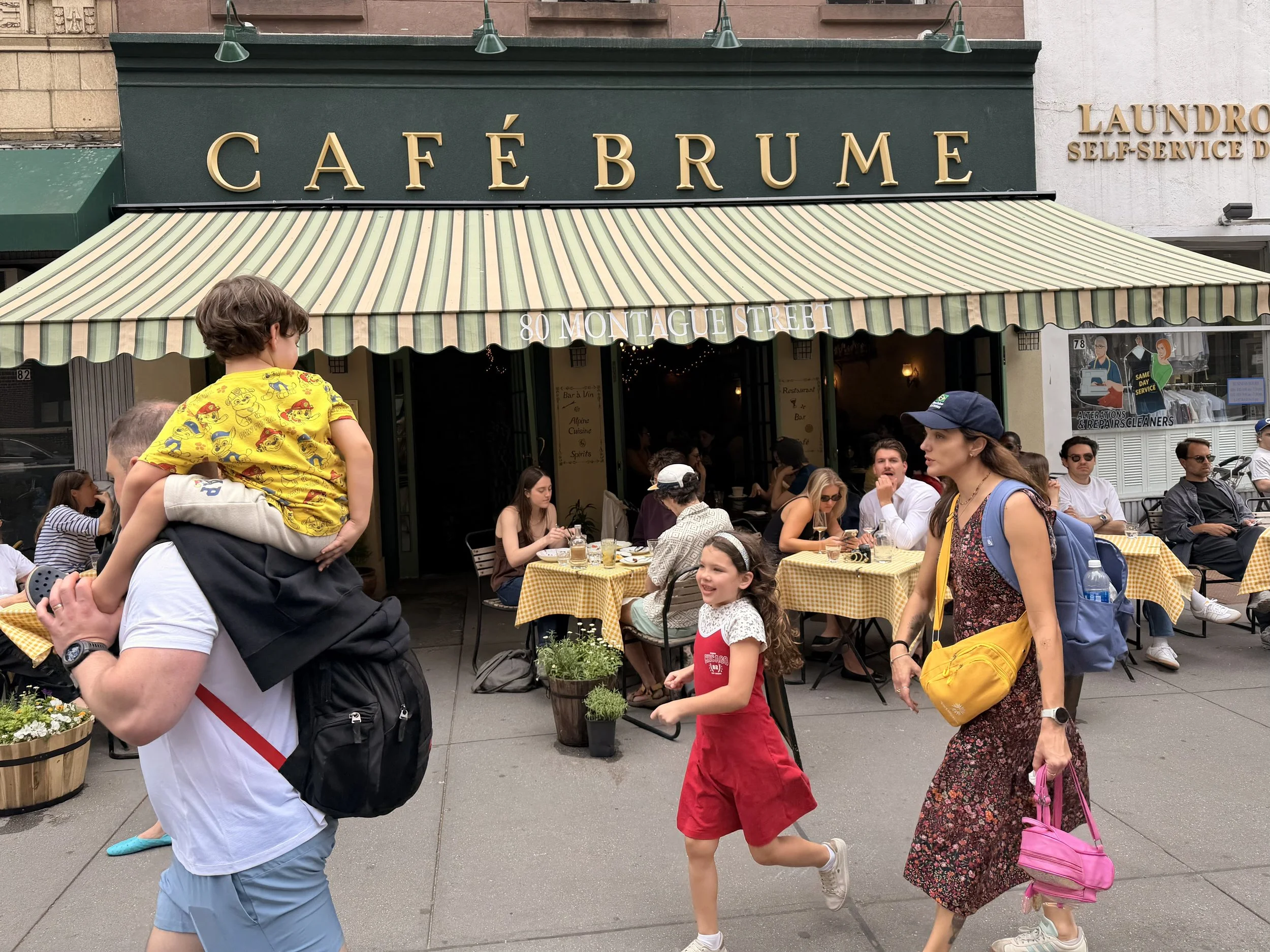 Outdoor patio scene outside Café Brume with people sitting at tables and walking along the sidewalk. The café has a green awning and gold lettering on the sign. 