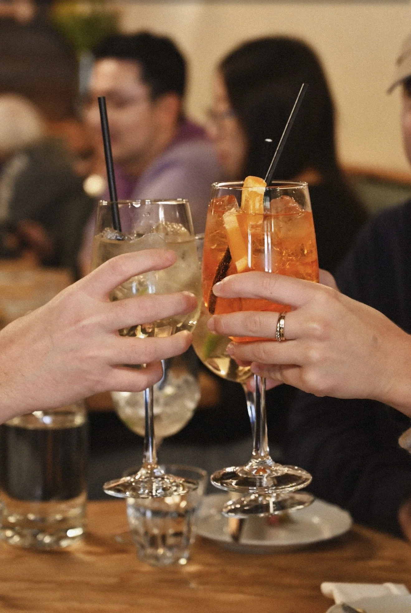 Two people are clinking glasses with spritz drinks, one is an Apénglow Fizz and the other is a Capelletti Spritz, during a social gathering indoors.