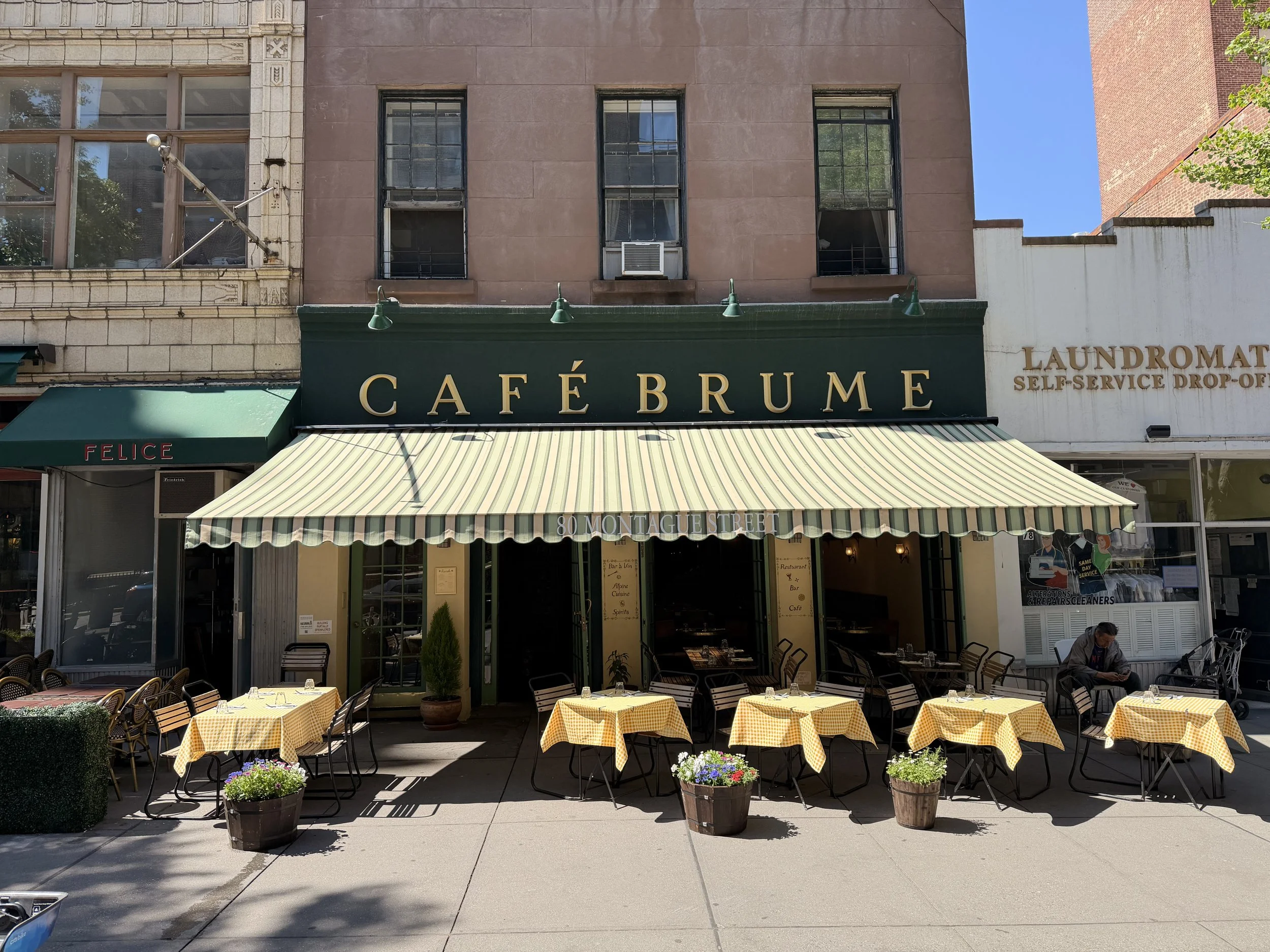Outdoor patio with yellow checkered tablecloths, chairs, potted plants, and a storefront with awning for Café Brûme at 80 Montague Street.