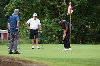 Four men playing golf on a green, with one preparing to putt near the flagstick, surrounded by trees.