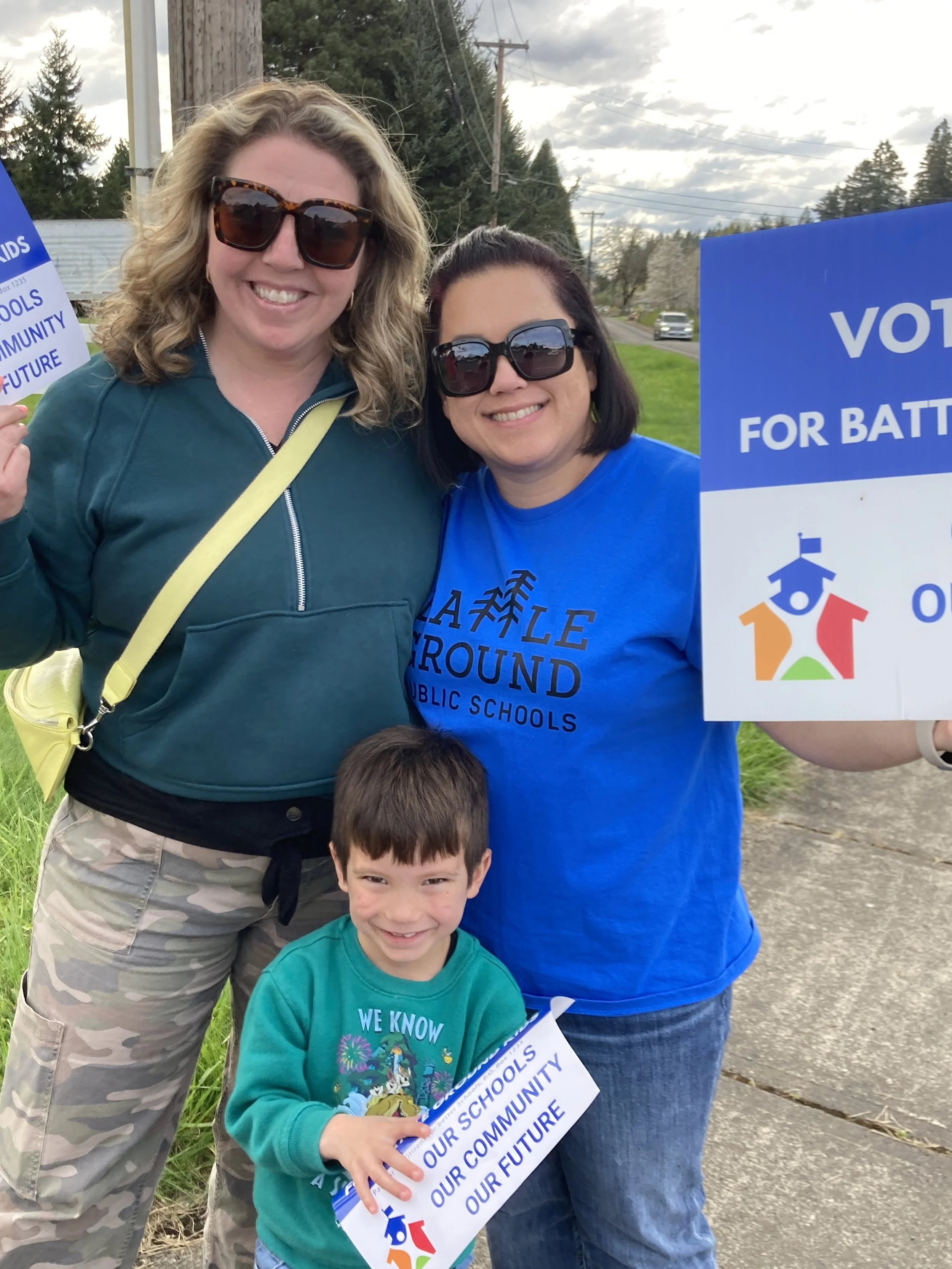 Two women and a young boy holding signs at an outdoor event, with the women wearing sunglasses and casual clothing, smiling, in a grassy area with trees and a street in the background, during a cloudy day.