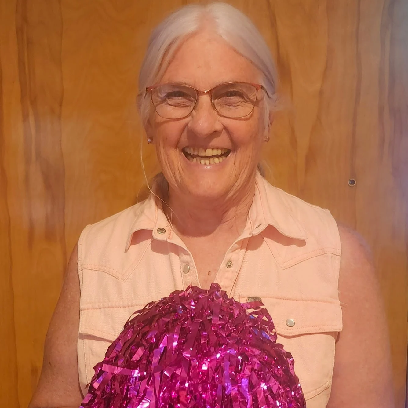 A woman with gray hair, glasses, and a big smile, holding a pink pom-pom, standing in front of a wooden wall.