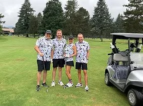 Four men in matching floral shirts standing on a golf course next to a golf cart.