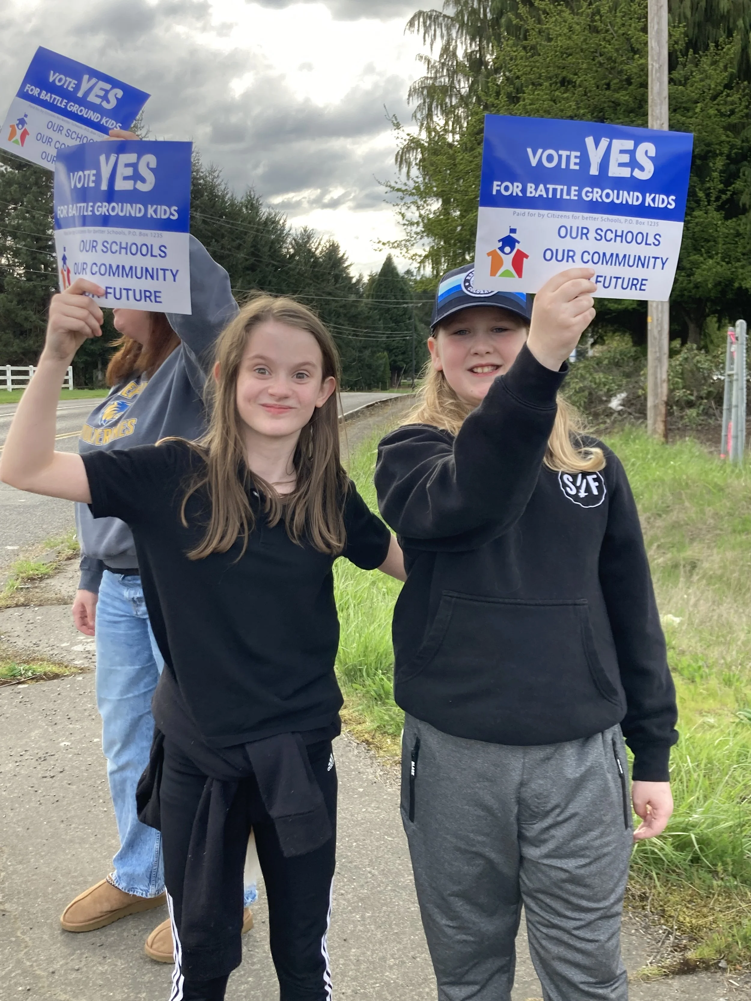 Two girls holding signs that read ‘Vote Yes for Battle Ground Kids’ and ‘Our Schools, Our Community, Our Future,’ standing outdoors on a sidewalk, with trees and a cloudy sky in the background.