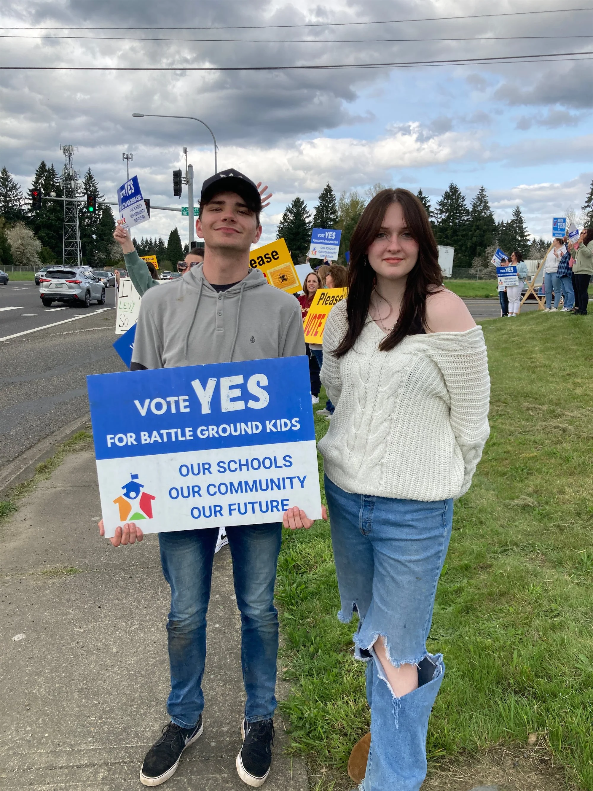 Two young people stand outdoors holding signs that support voting yes for a measure related to battle ground kids. The young man on the left wears a gray hoodie and black sneakers, and the young woman on the right wears a white off-the-shoulder sweater and ripped jeans. In the background, several other people hold similar signs; the sky is partly cloudy.
