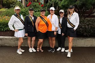 Six women standing outdoors near a low stone wall, dressed in white and black with some wearing orange, all wearing hats and sunglasses.