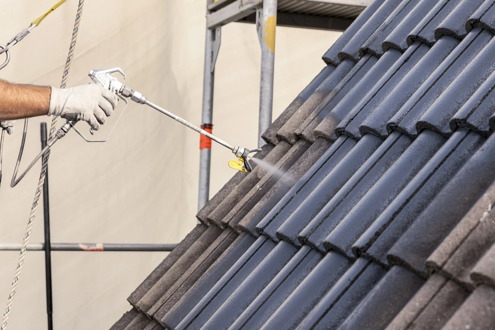 Ein Handwerker sprüht eine Beschichtung auf Dachziegel mit einem Airless-Gerät. Der Dachfirst besteht aus welligen Dachziegeln, und es ist eine Gerüstkonstruktion im Hintergrund sichtbar.
