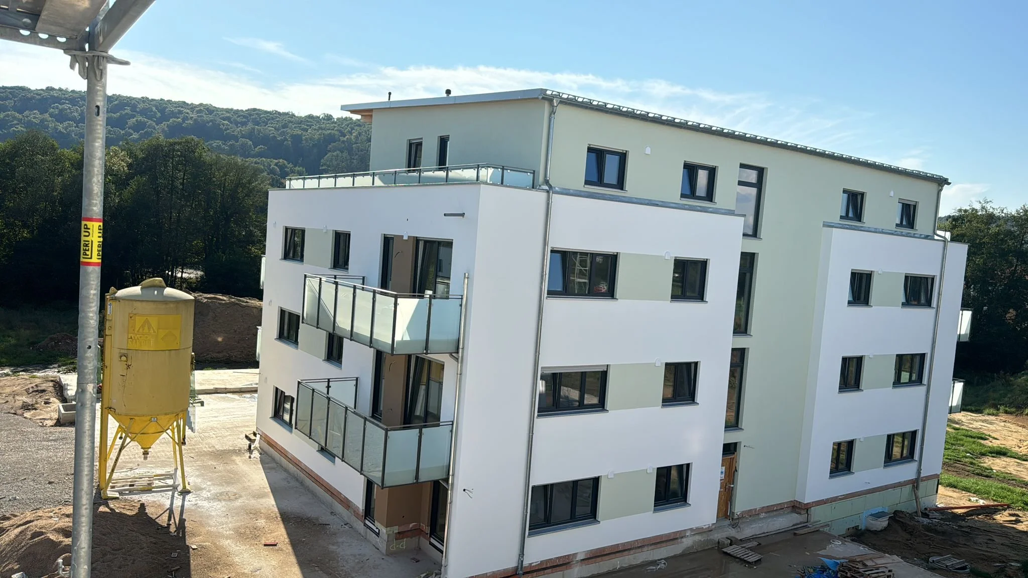 Neues mehrstöckiges Apartmentgebäude unter Bau, mit Balkonen, angrenzendem Bauplatz, Bauausrüstung und einer grünen, bewaldeten Hügel im Hintergrund bei sonnigem Wetter.