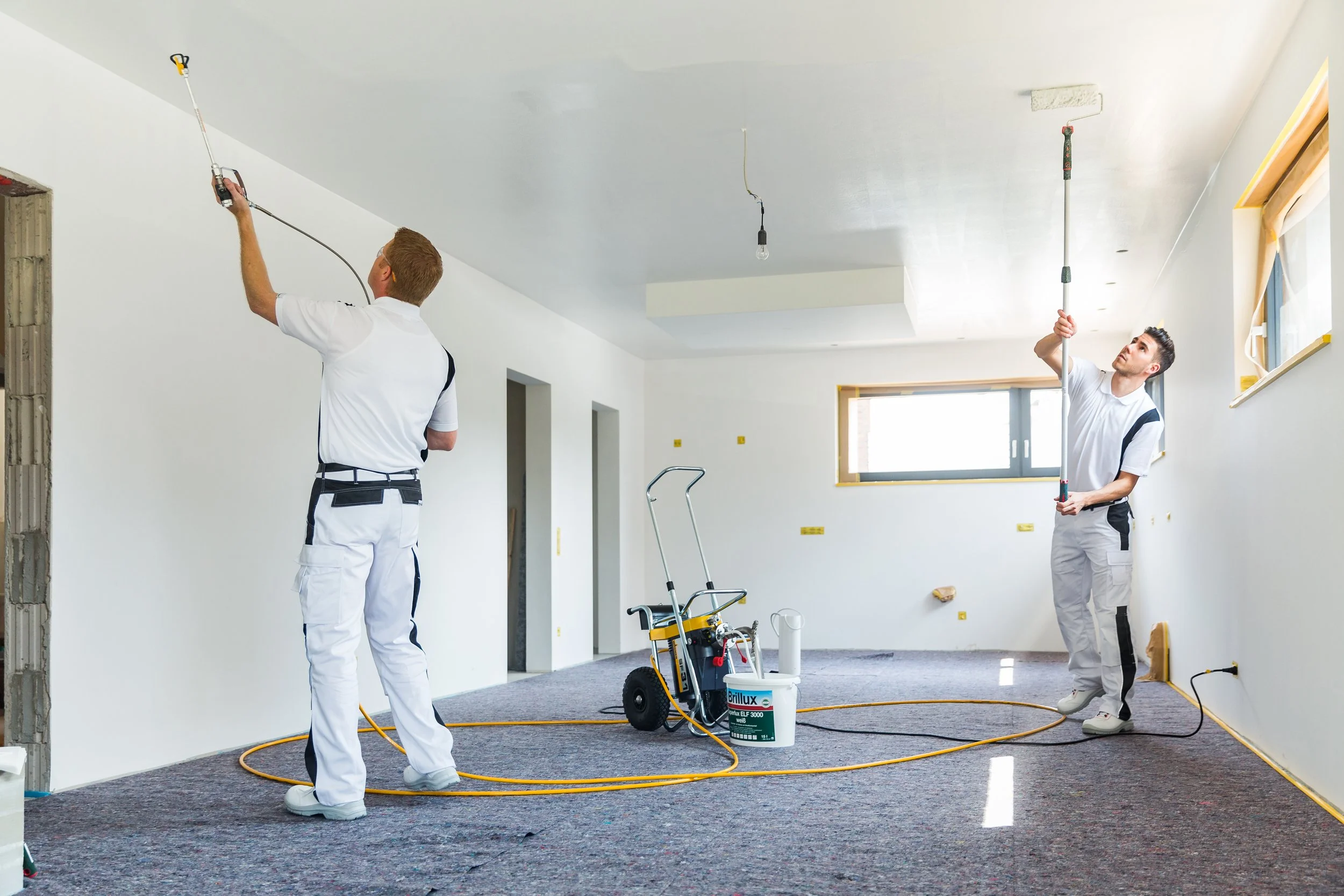Zwei Handwerker streichen die Decke in einem Raum, mithilfe eines Airless-Gerätes.