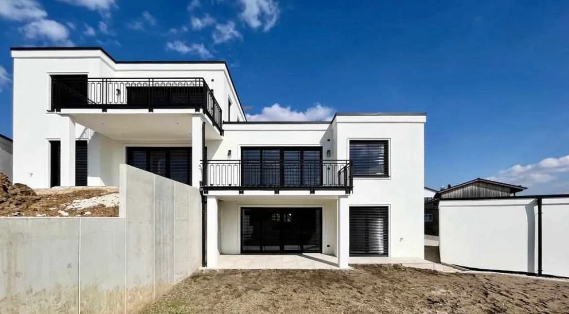Modernes weißes Haus mit mehreren Balkonen und schwarzen Fenstern unter blauem Himmel.