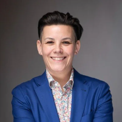 A woman with short dark hair wearing a blue blazer and a floral shirt, smiling with crossed arms in front of a gray background.