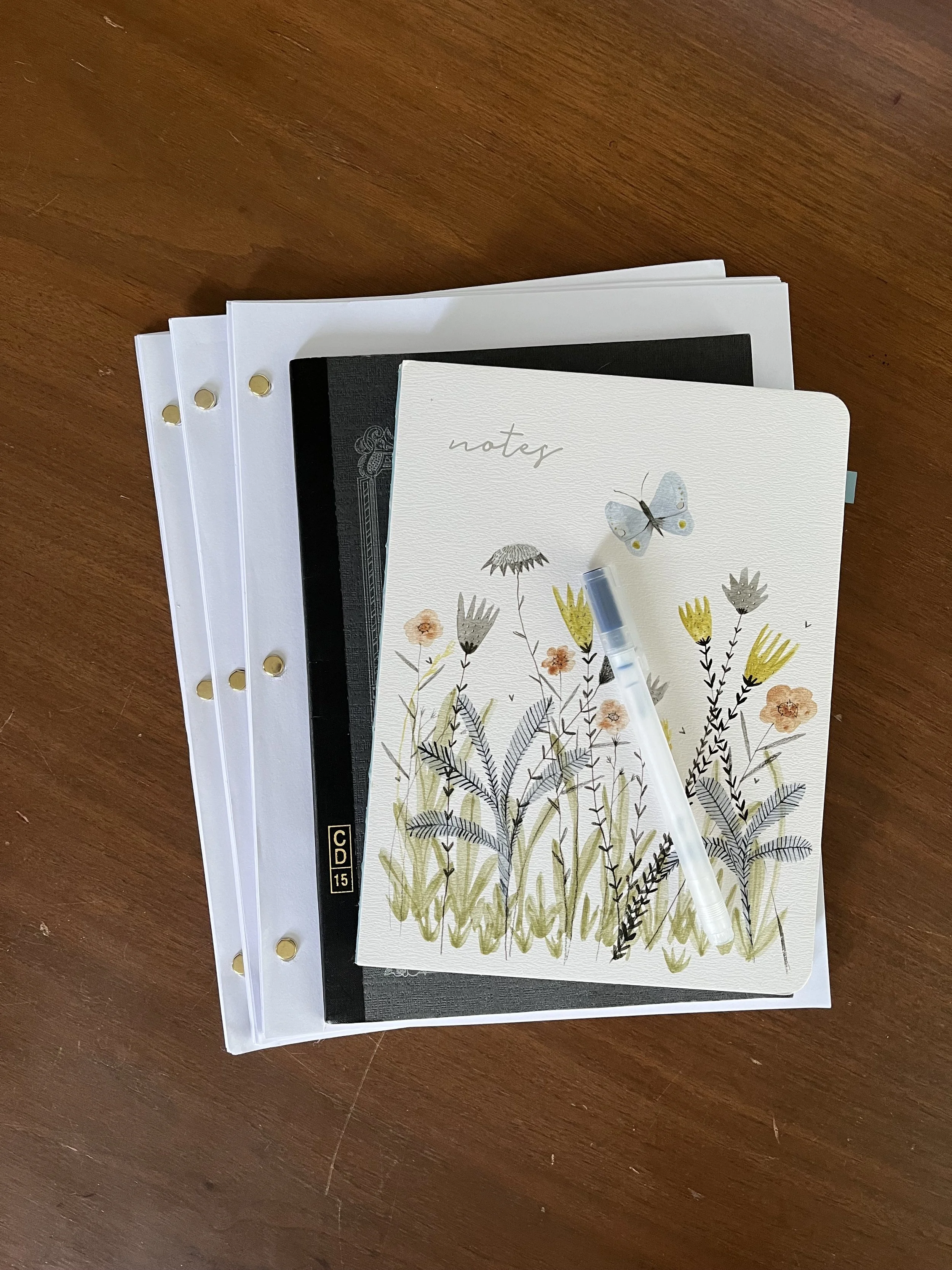 Stack of notebooks and papers on a wooden surface, featuring a notebook with floral and butterfly design on top.