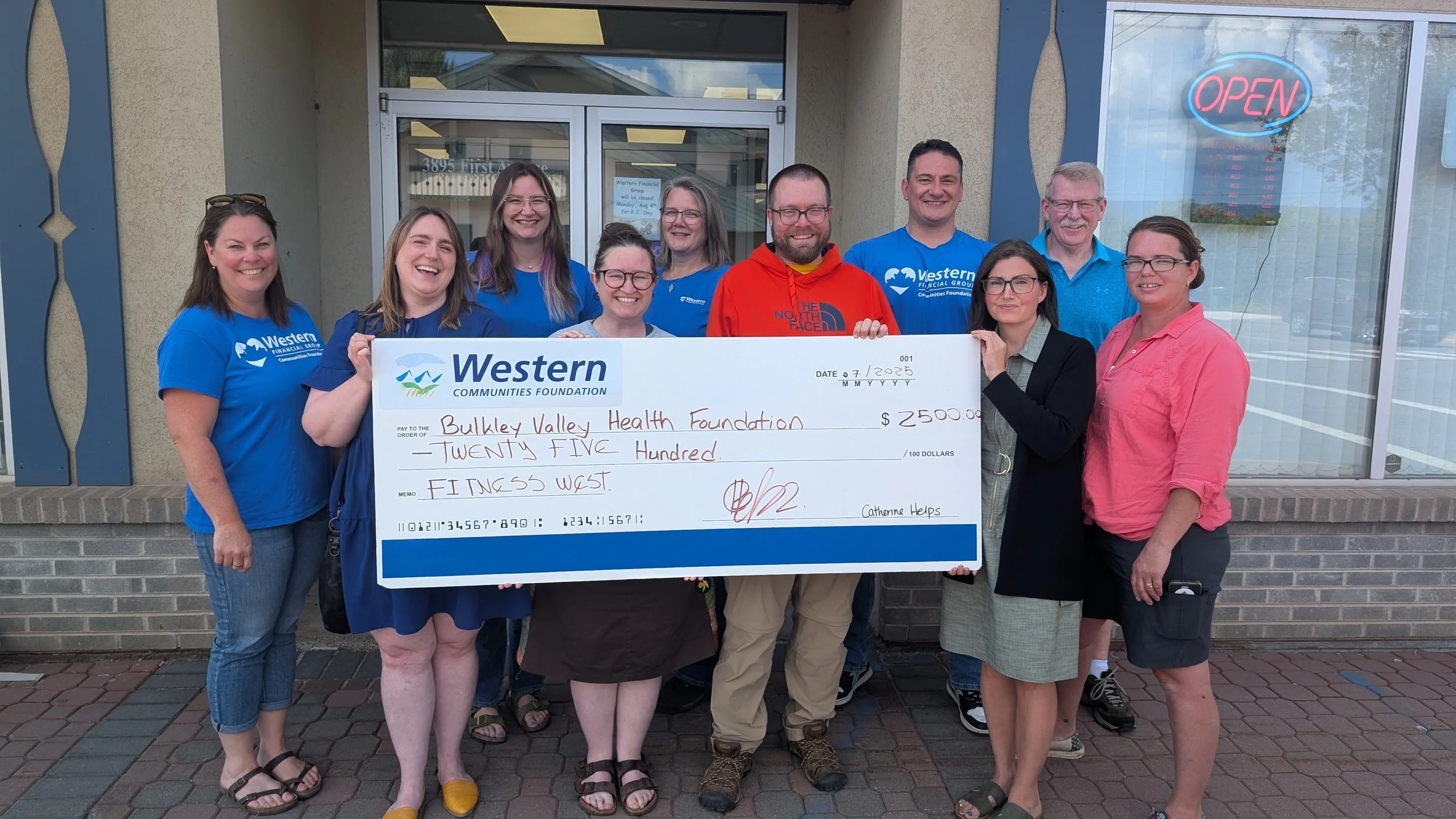 Group of ten people standing outside a building holding an oversized check for 2500 dollars made out to Buckley Valley Health Foundation, with a bright smiling woman in green and black standing on the right.