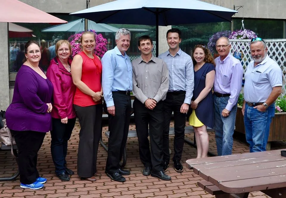A group of nine people standing outdoors under umbrellas, smiling for a photo.