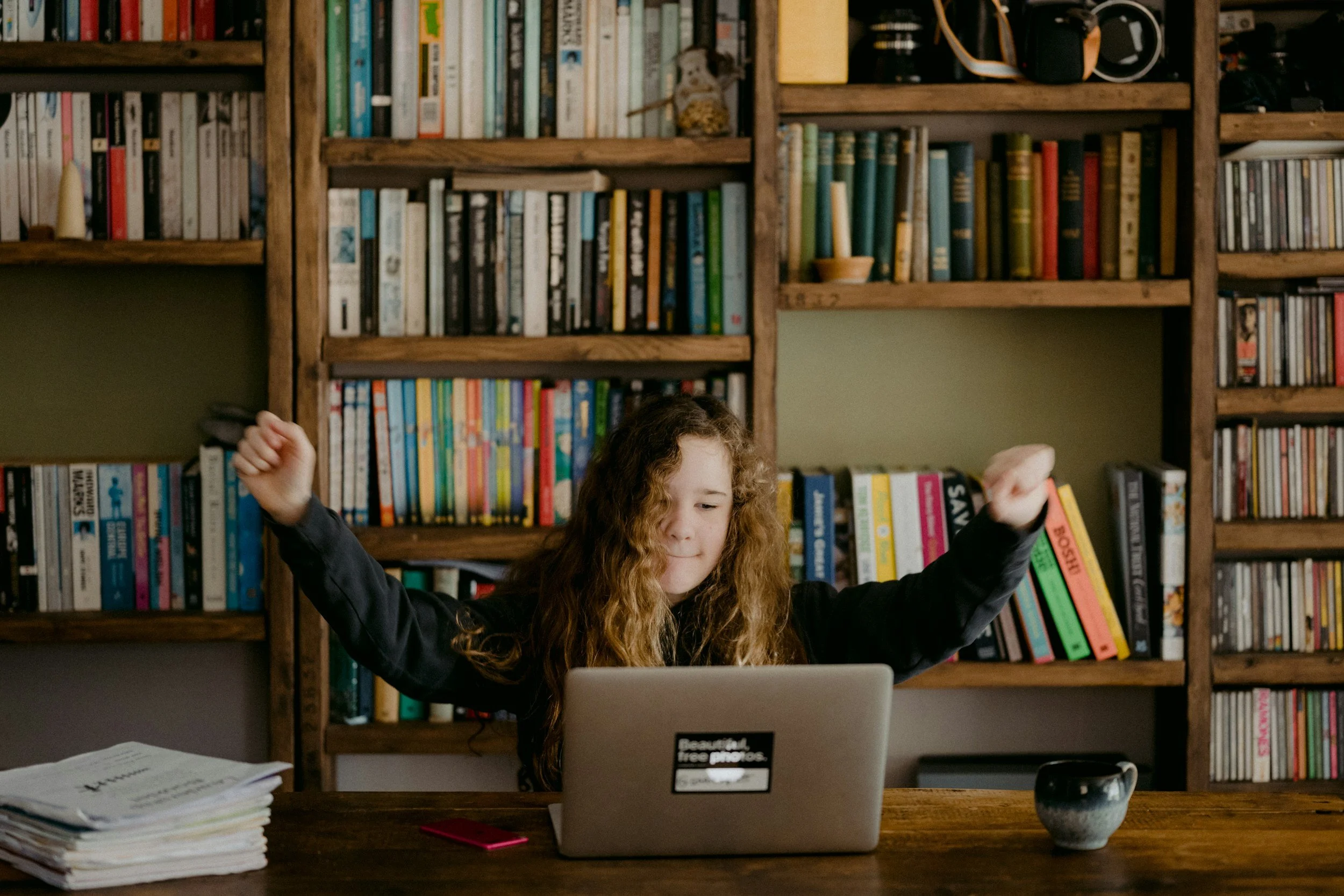 A young girl with long curly hair sitting at a wooden desk in front of a laptop, raising her fists in a celebratory gesture, with a large bookshelf filled with books and a camera in the background.