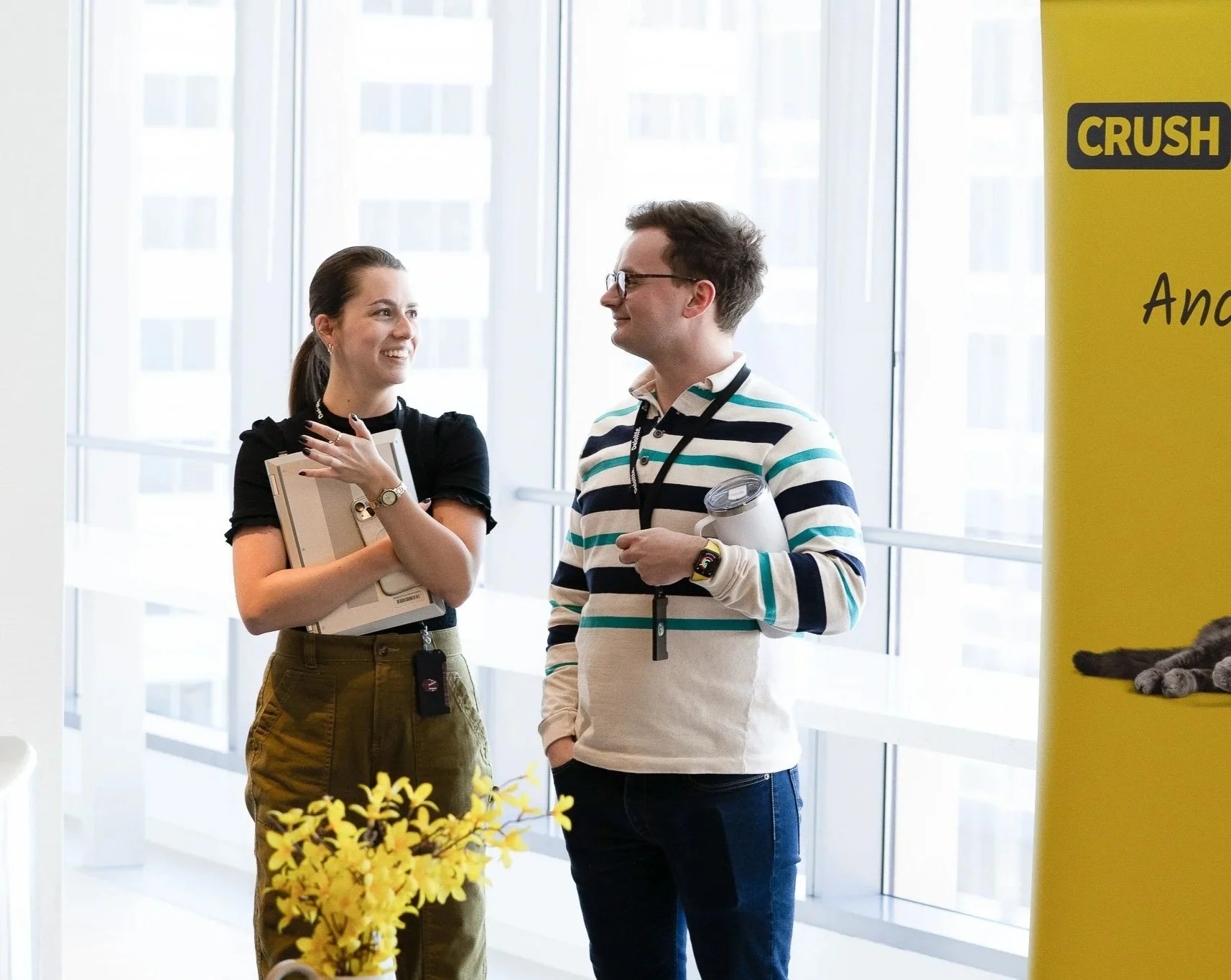 Two people are having a conversation in a bright, modern office with large windows. The woman on the left is holding a laptop and smiling, while the man on the right is wearing glasses and a striped sweater, holding a water bottle. There's a yellow s