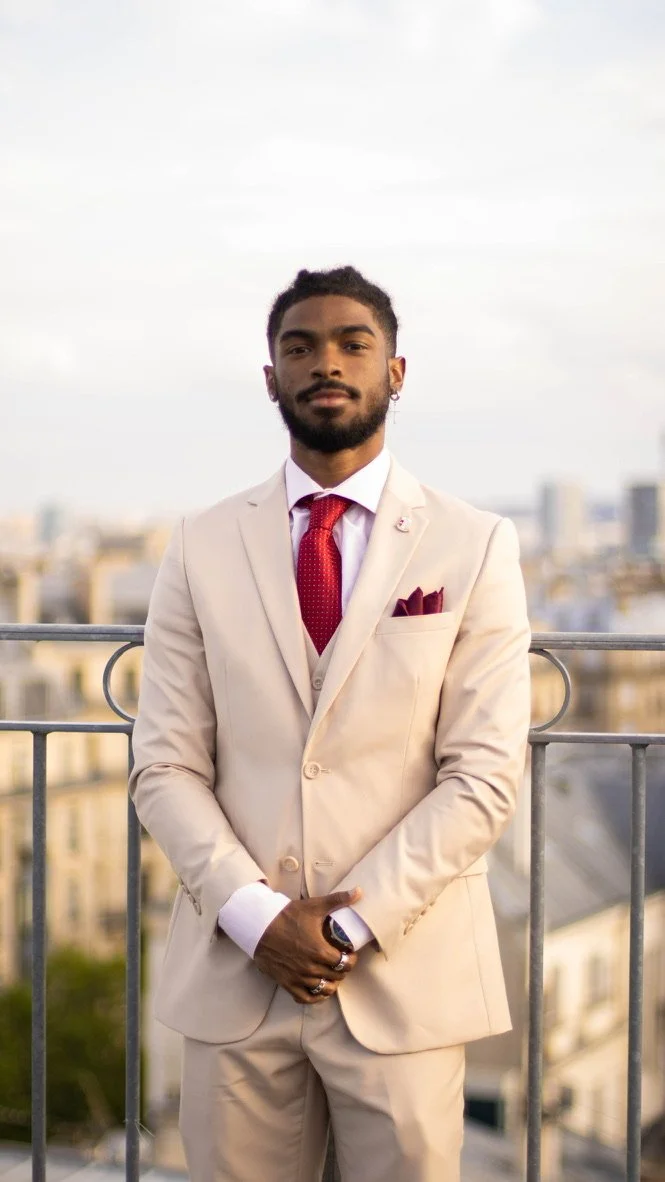 Man in beige suit with red tie and pocket square standing in front of a railing with cityscape background.