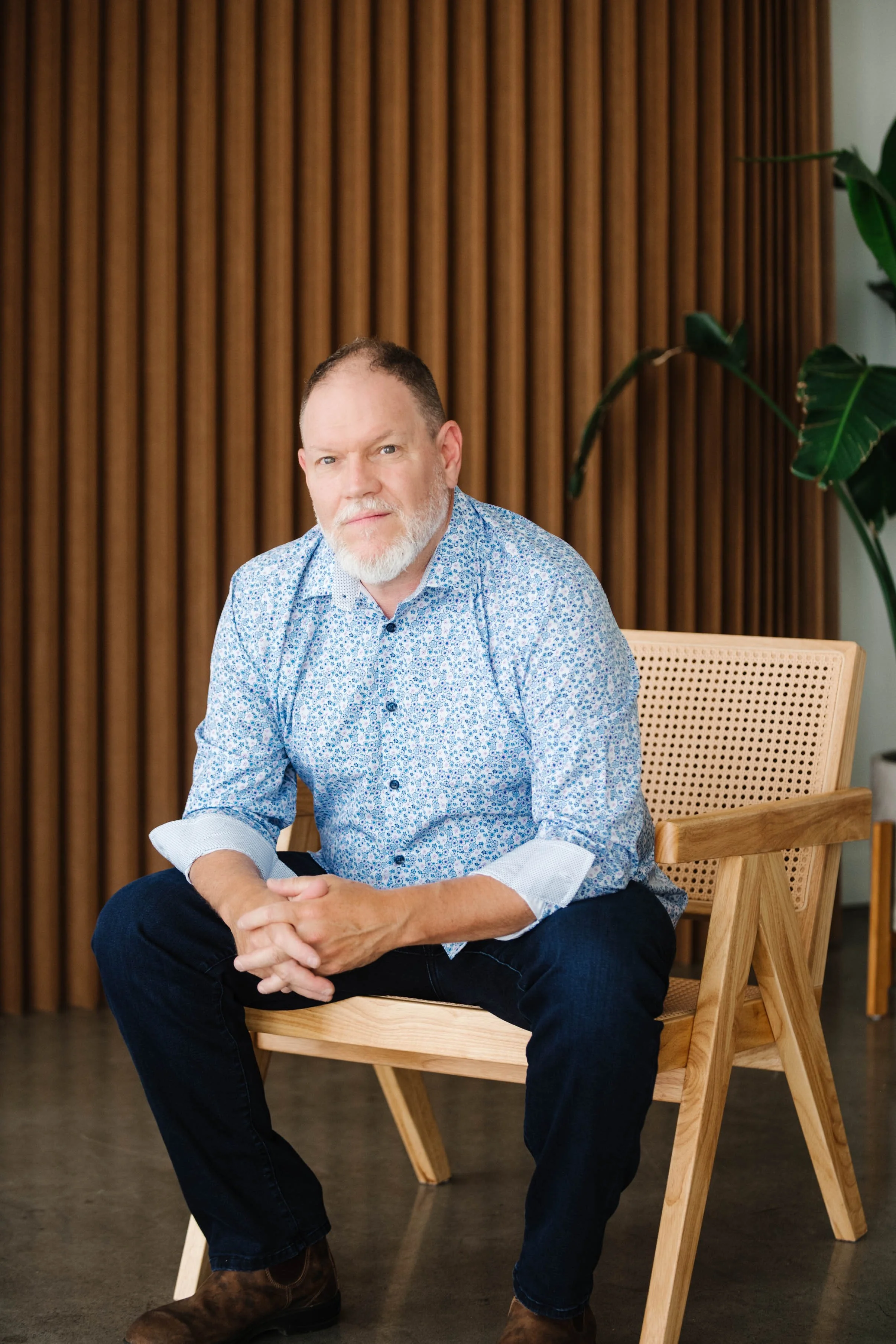 A man with a beard and light hair, wearing a blue patterned shirt, sits on a wooden chair with a rattan backrest, in front of a wooden slat wall and next to a large green plant.