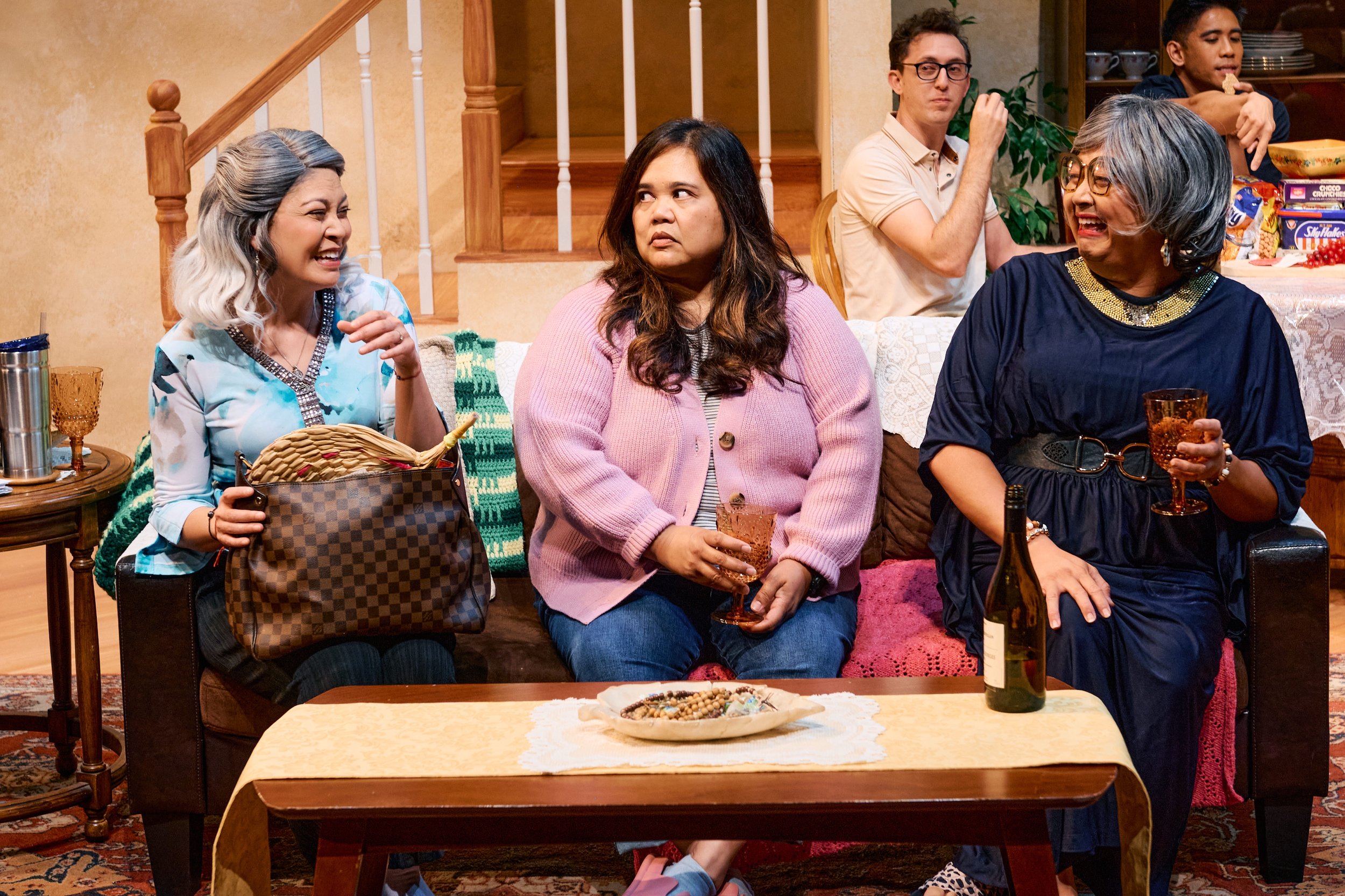 A group of people in a cozy living room, sitting on a couch. Two women on the left and right are smiling and holding drinks, while the woman in the center looks thoughtful. There is a wooden coffee table with snacks and a wine bottle in front of them