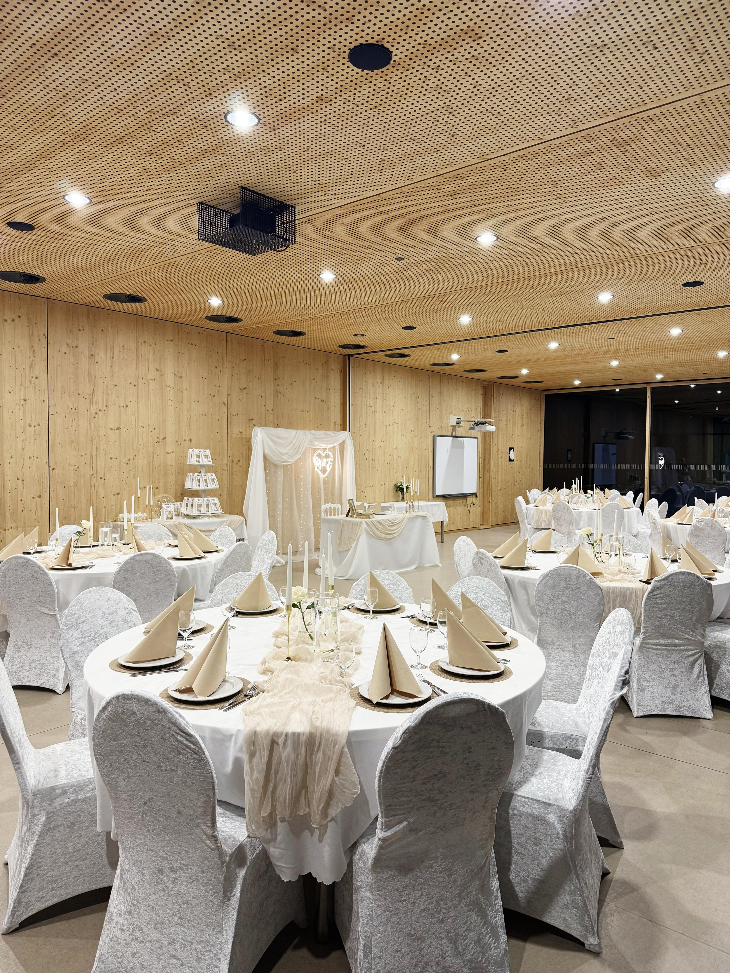 Ein festlich gedeckter Veranstaltungsraum mit runden Tischen, weißen Stuhlhussen, Kerzen, Besteck und Wein gläsern, sowie einer dekorativen Bühne mit weißen Drapierungen und Herzleuchte.
