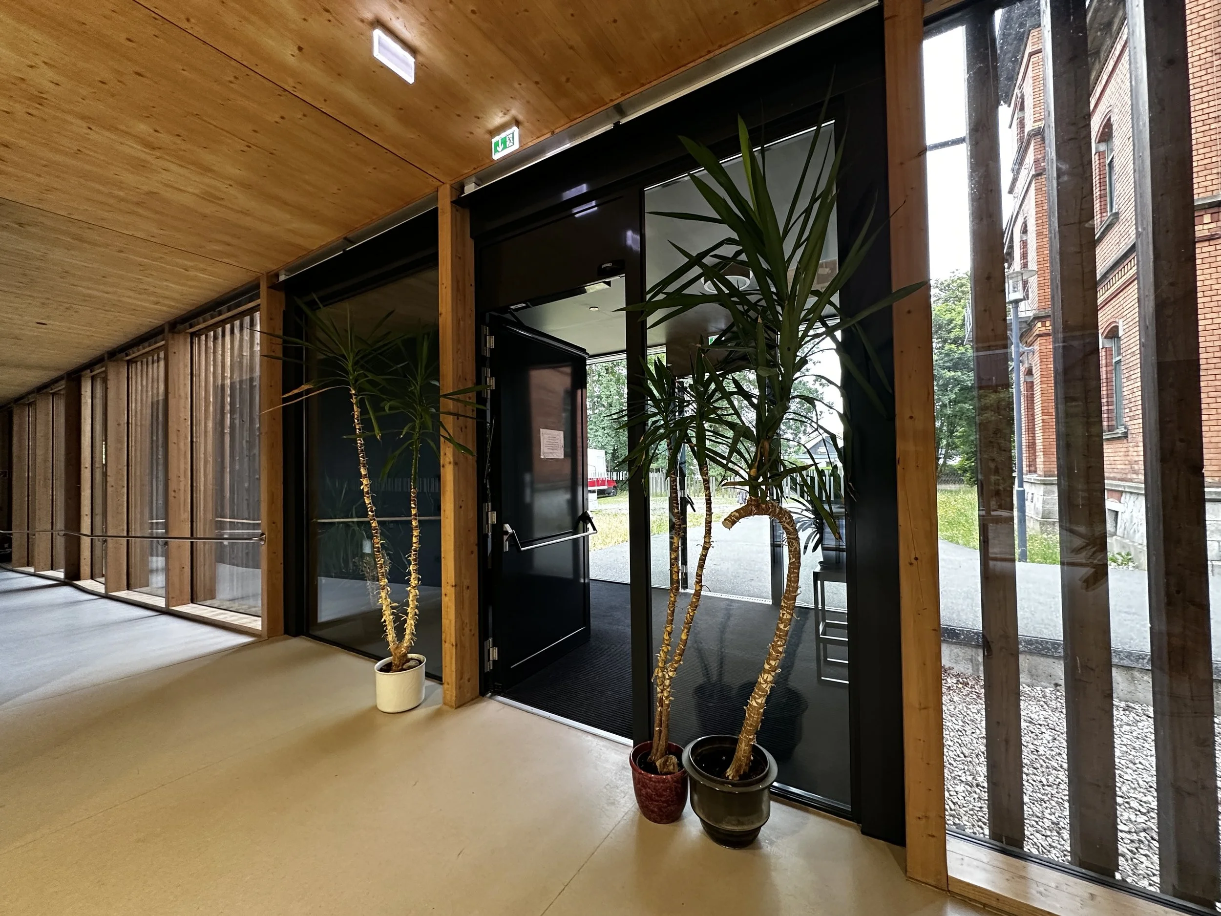 Innenaufnahme eines Gebäudes mit Holzwänden und großen Fenstern, zwei Topfpflanzen stehen vor einem schwarzen Eingangstür.