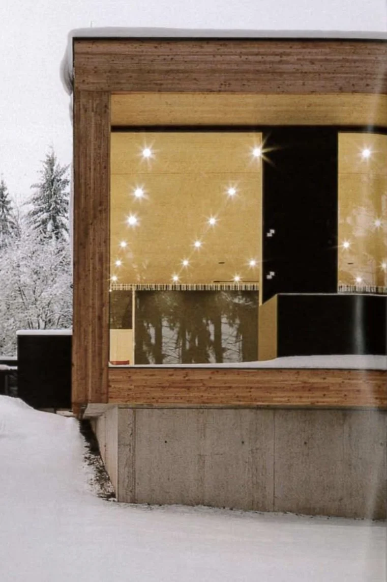 Das Bild zeigt eine moderne Holzwand mit großen Fenstern, durch die man nach innen in eine gut beleuchtete, stilvolle Raumfläche mit Holzpaneelen und Reflexionen von Bäumen im Hintergrund sehen kann. Draußen liegt Schnee.