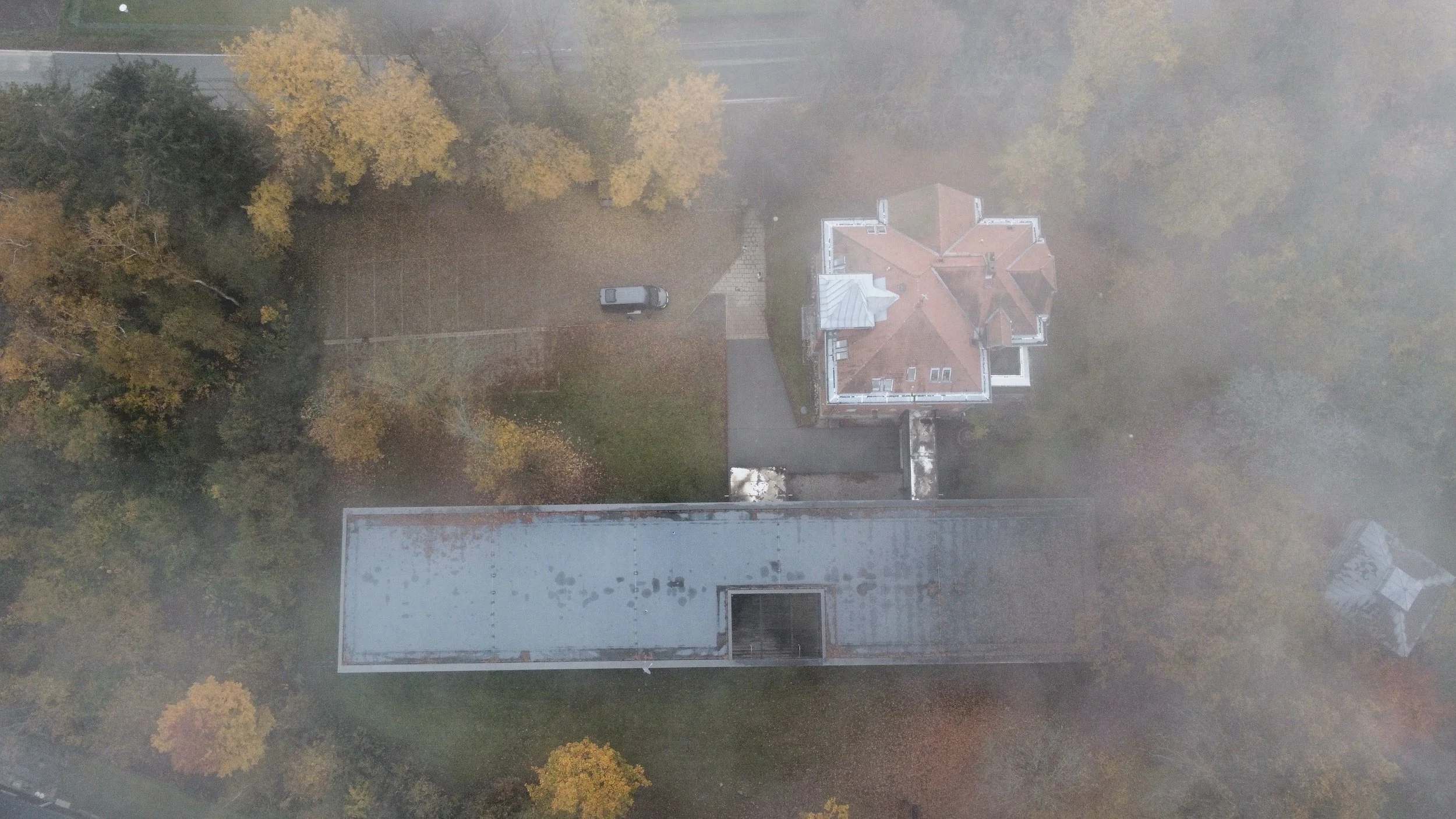 Vogelperspektive auf ein Flachdach eines Gebäudes mit rötlichem Ziegeldach, umgeben von Bäumen mit Herbstlaub, und einem kleinen Parkplatz mit einem Fahrzeug.