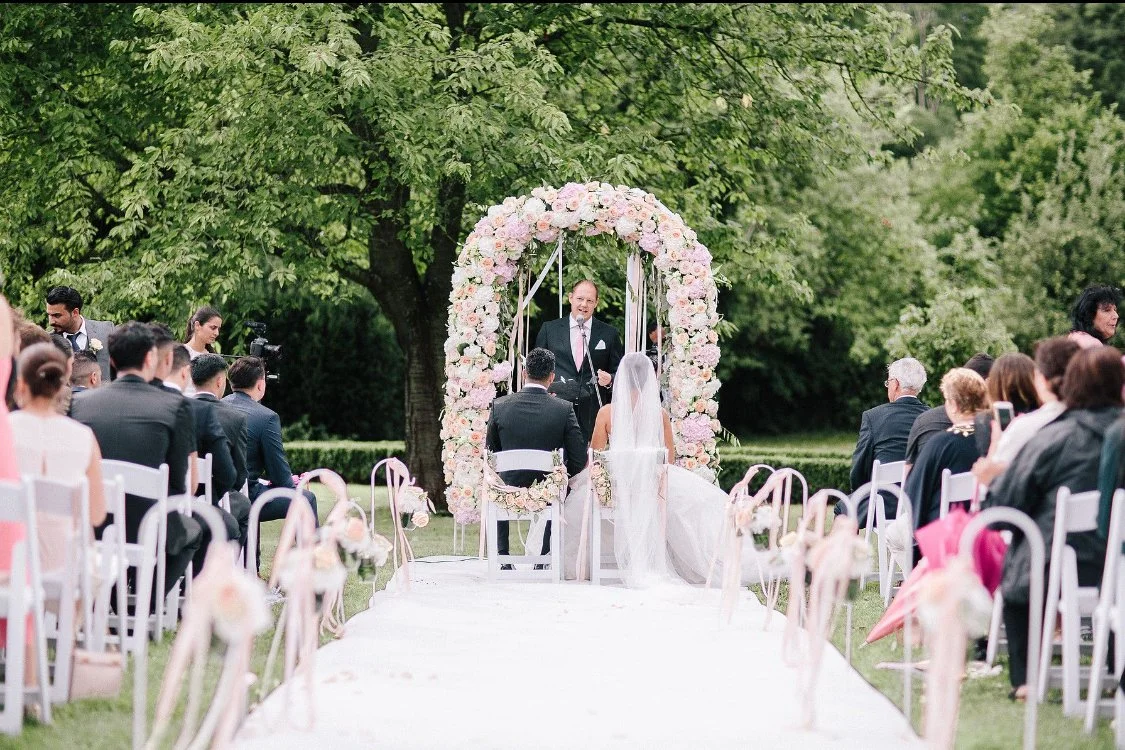 Hochzeitszeremonie im Freien mit Braut, Bräutigam und Gästen unter einem blumengeschmückten Bogen, umgeben von Bäumen.