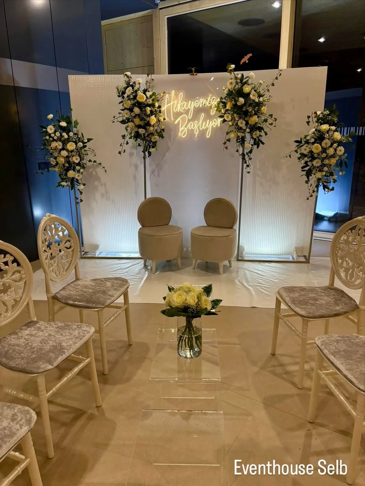 Ein Bereich mit two beige Stühle vor einem weißen Hintergrund und blumenartigen Dekorationen. Es gibt eine Neon-Schrift, die sagt 'Hayatimer Başlayor'.