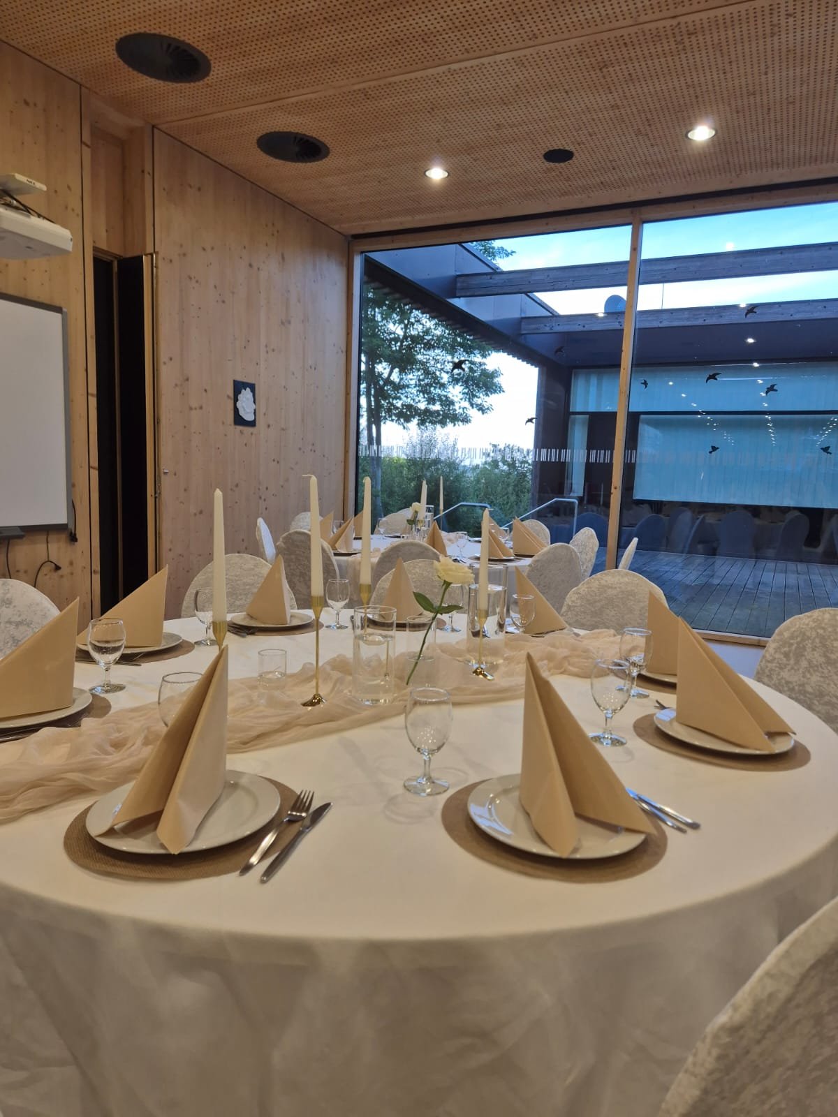 Festlich gedeckter Esstisch mit weißen Servietten, Kerzen, Wasser- und Weingläsern in einem modernen Raum mit Holzwänden und großen Fenstern
