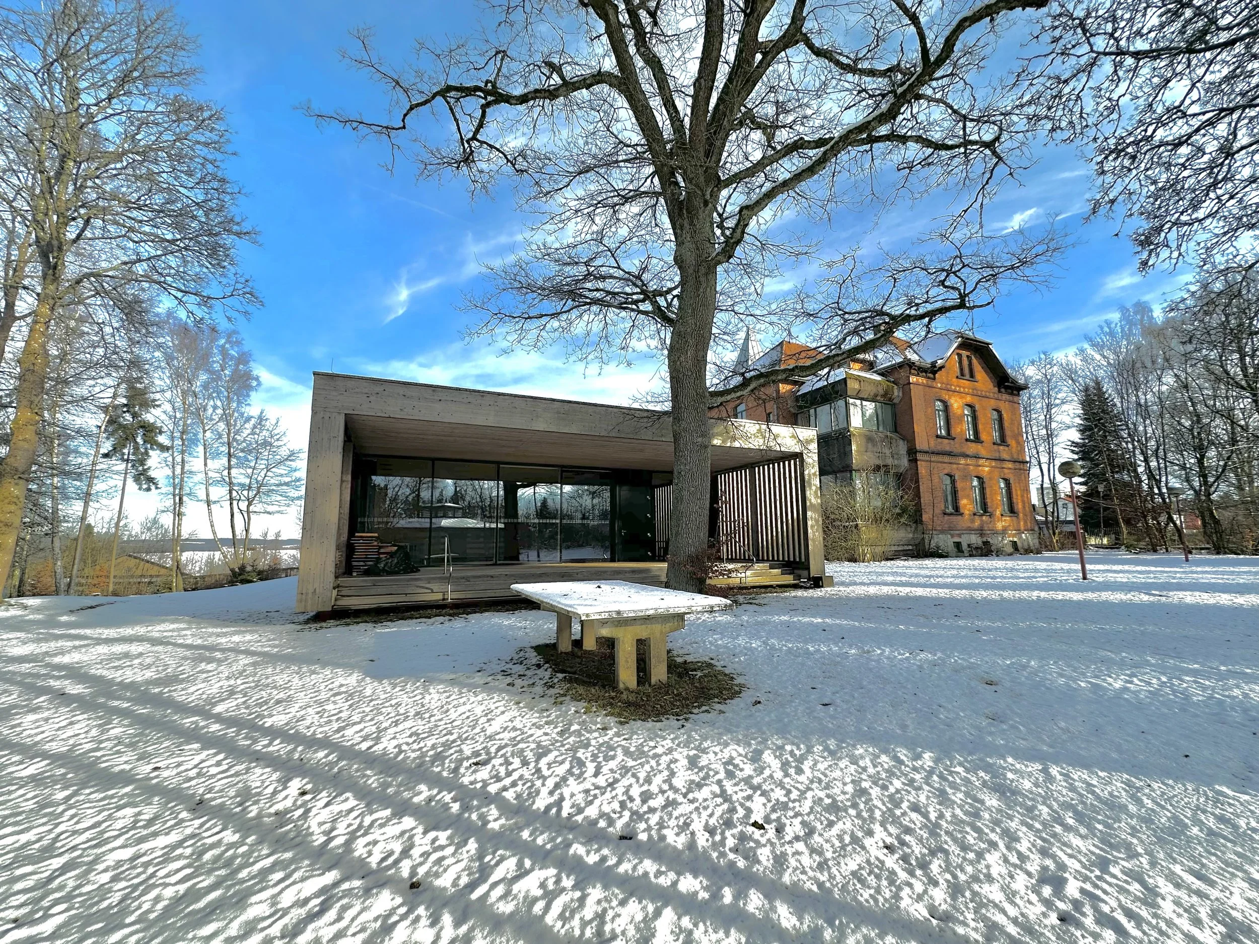 Moderne Holz- und Glaskonstruktion in winterlicher Landschaft mit Schnee bedecktem Boden, großen Bäumen und einem roten Backsteingebäude im Hintergrund.