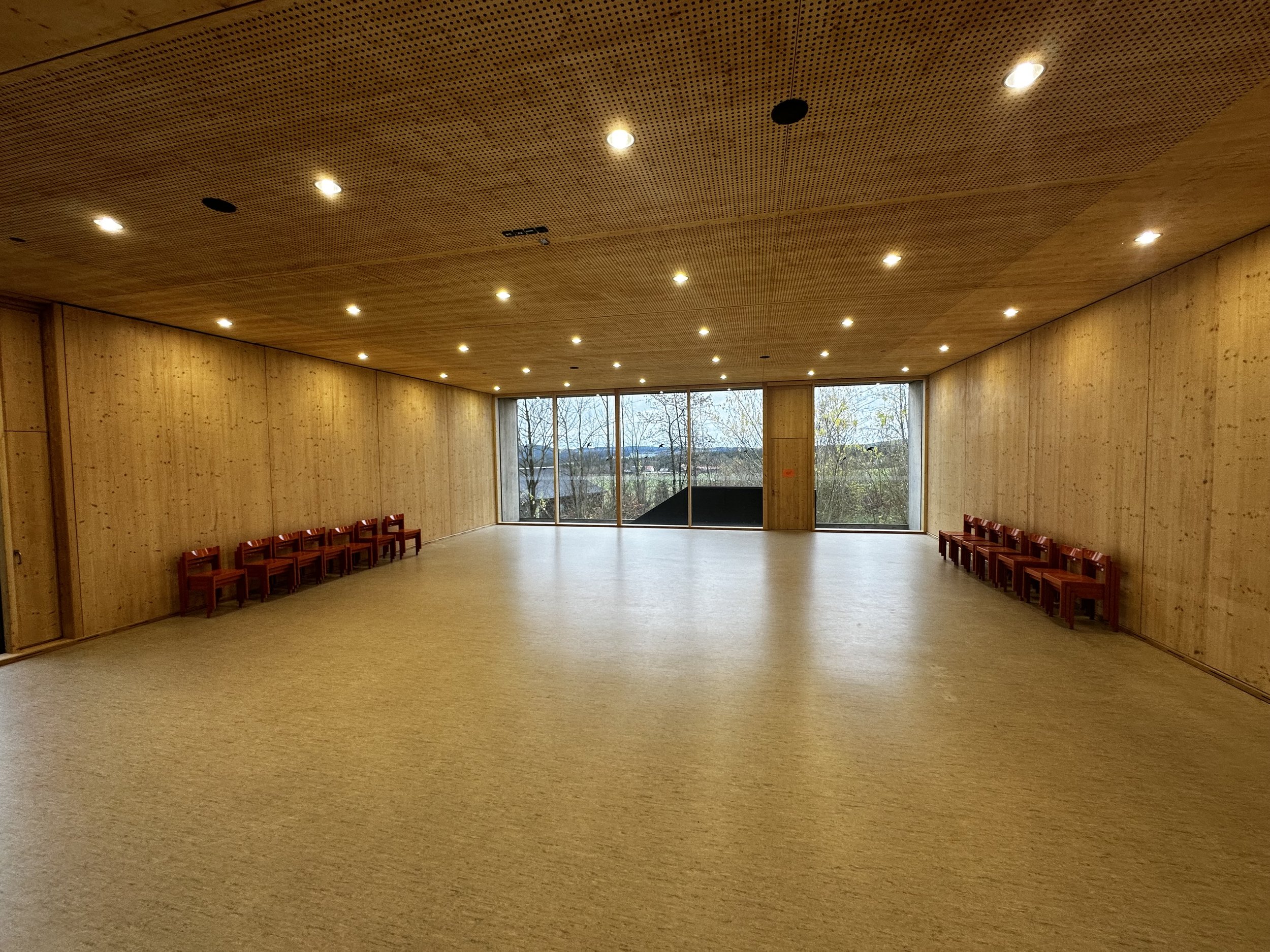 Leeres, helles Holzsaal mit Sitzbänken an den Wänden und großen Fenstern im Hintergrund, die einen Blick nach draußen ermöglichen.