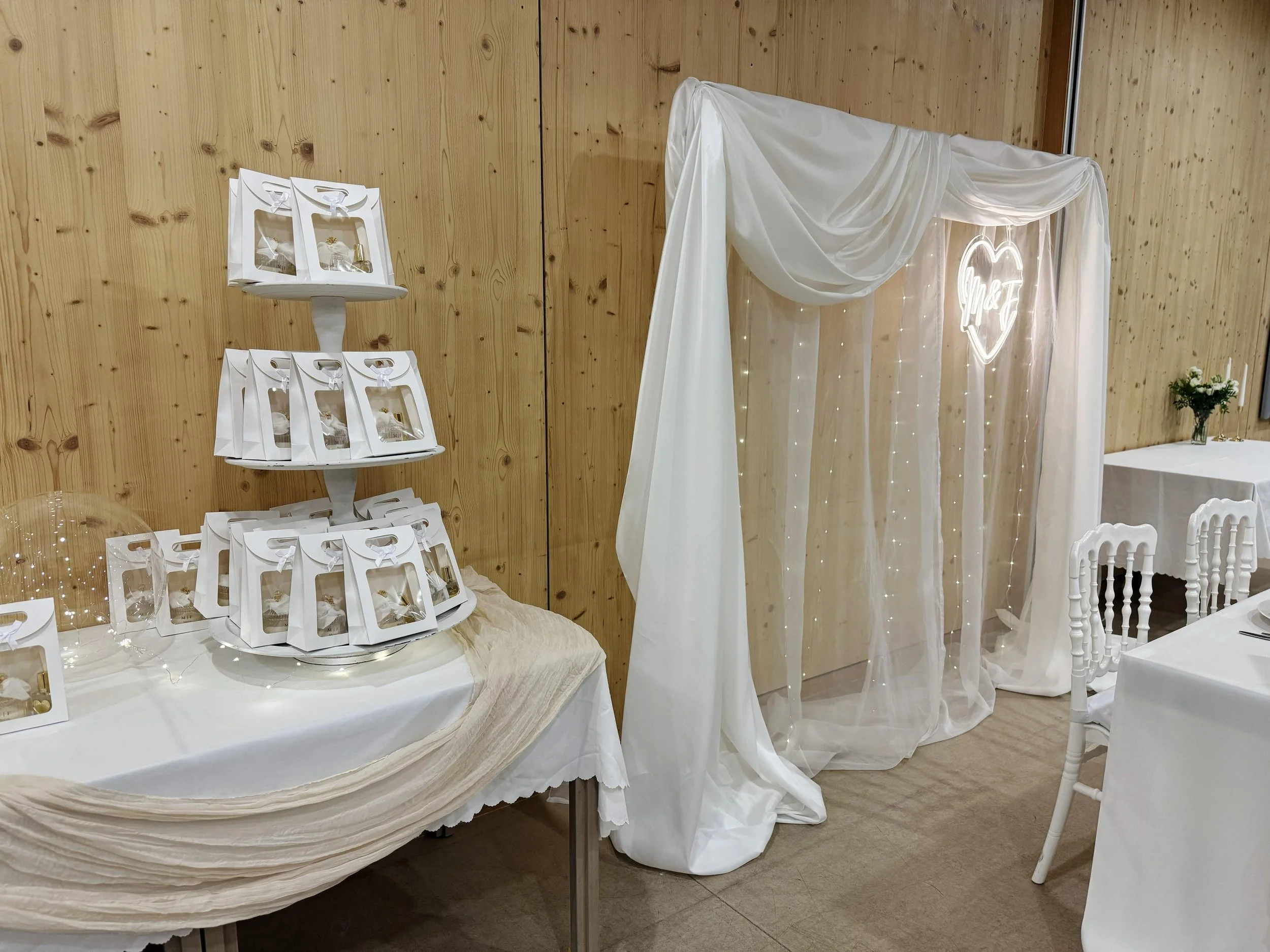 Weiße Hochzeitsdeko mit Herzmotiv und Lichterkette, elegante Tische, kleine Geschenkkästen, Blumendeko auf Holzwand