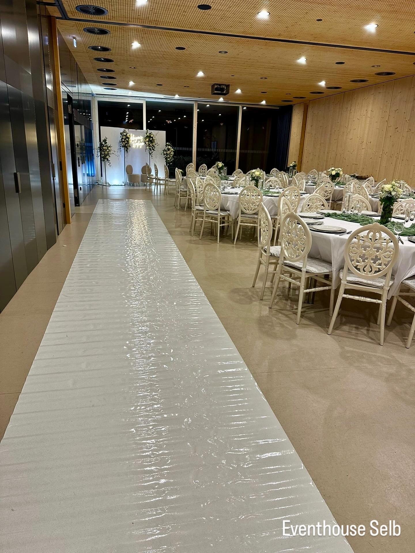 Ein festlich gedeckter Veranstaltungsraum mit weißen Stühlen und Tischen, Dekorationen mit Blumen, im Hintergrund eine weiße Wand mit Blumenarrangements, im vorderen Bereich eine weiße, gepolsterte Laufbahn.