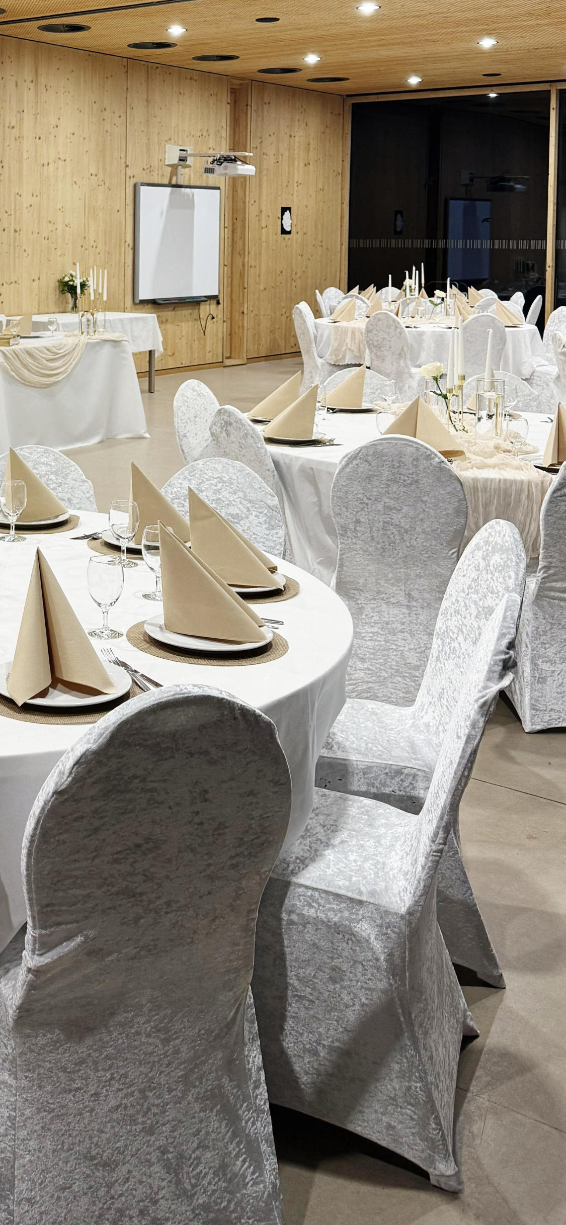 Eleganter festlich gedeckter Raum mit runden Tischen, weißem Tischtuch, beige Stoffservietten, edlem Geschirr, Gläsern und Kerzen, bereit für eine Feier oder Hochzeitsveranstaltung.