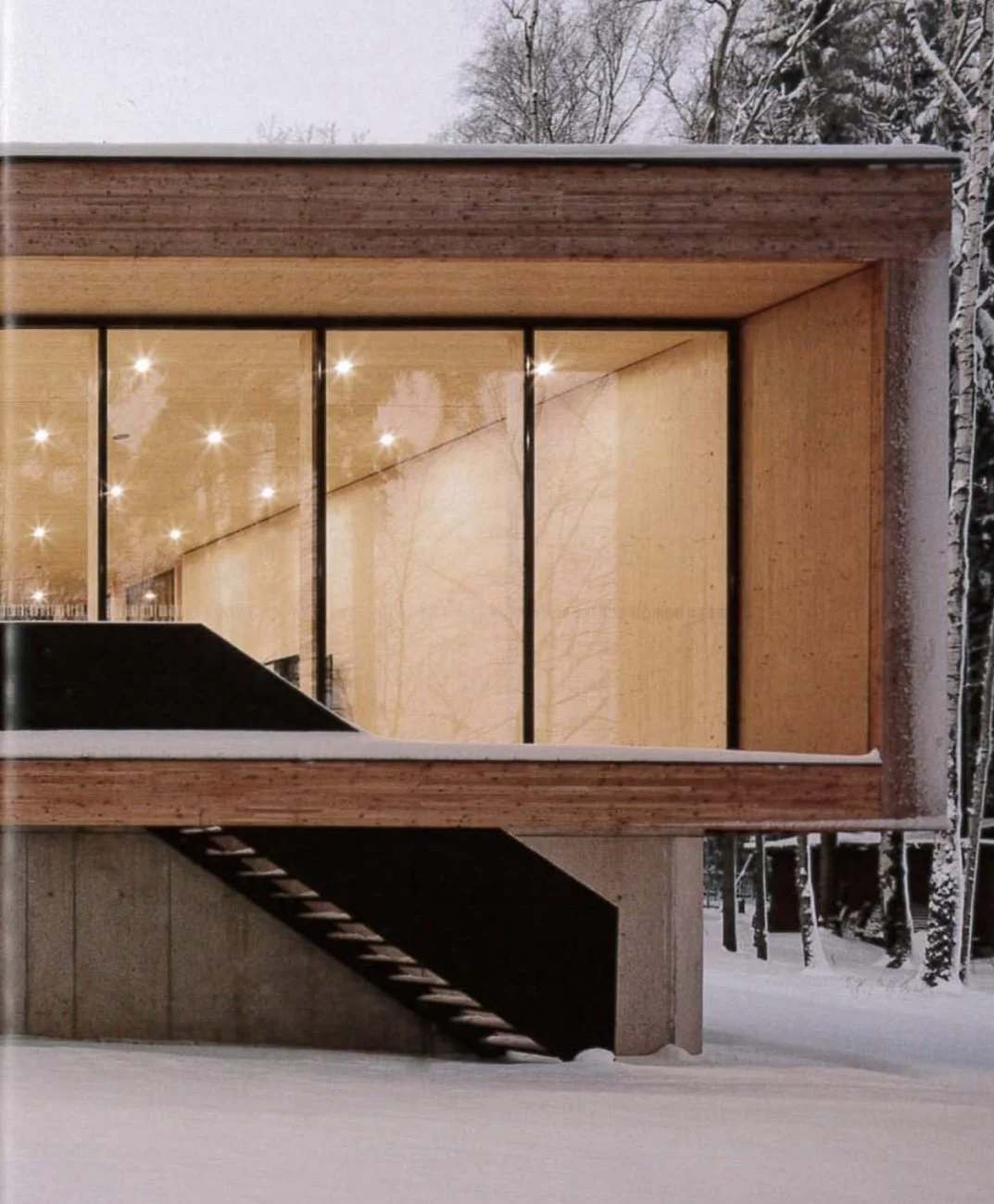 Modernes Holzhaus im Winter bei Nacht mit beleuchtetem Innenraum, sichtbaren Treppen und großen Fenstern, umgeben von Schneelandschaft und Bäumen.