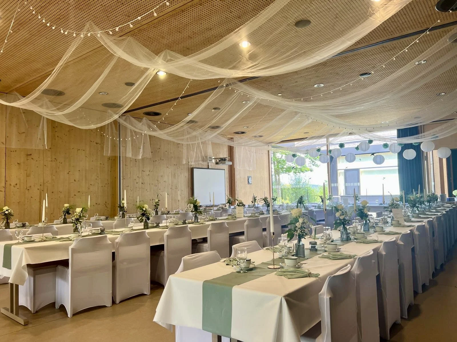 Ein festlich gedeckter Raum mit langen Tischen, weißen Tischdecken, Blumenvasen, Kerzen, Geschirr und Besteck, dekoriert mit weißen Papierlaternen und Stoffrosetten an der Decke, im Hintergrund große Fenster mit Blick ins Freie.