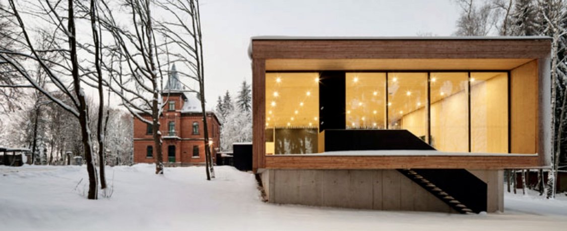 Moderne Holzkonstruktion mit großen Glasfenstern in winterlicher Schneelandschaft, hinter alten roten Backsteingebäude.