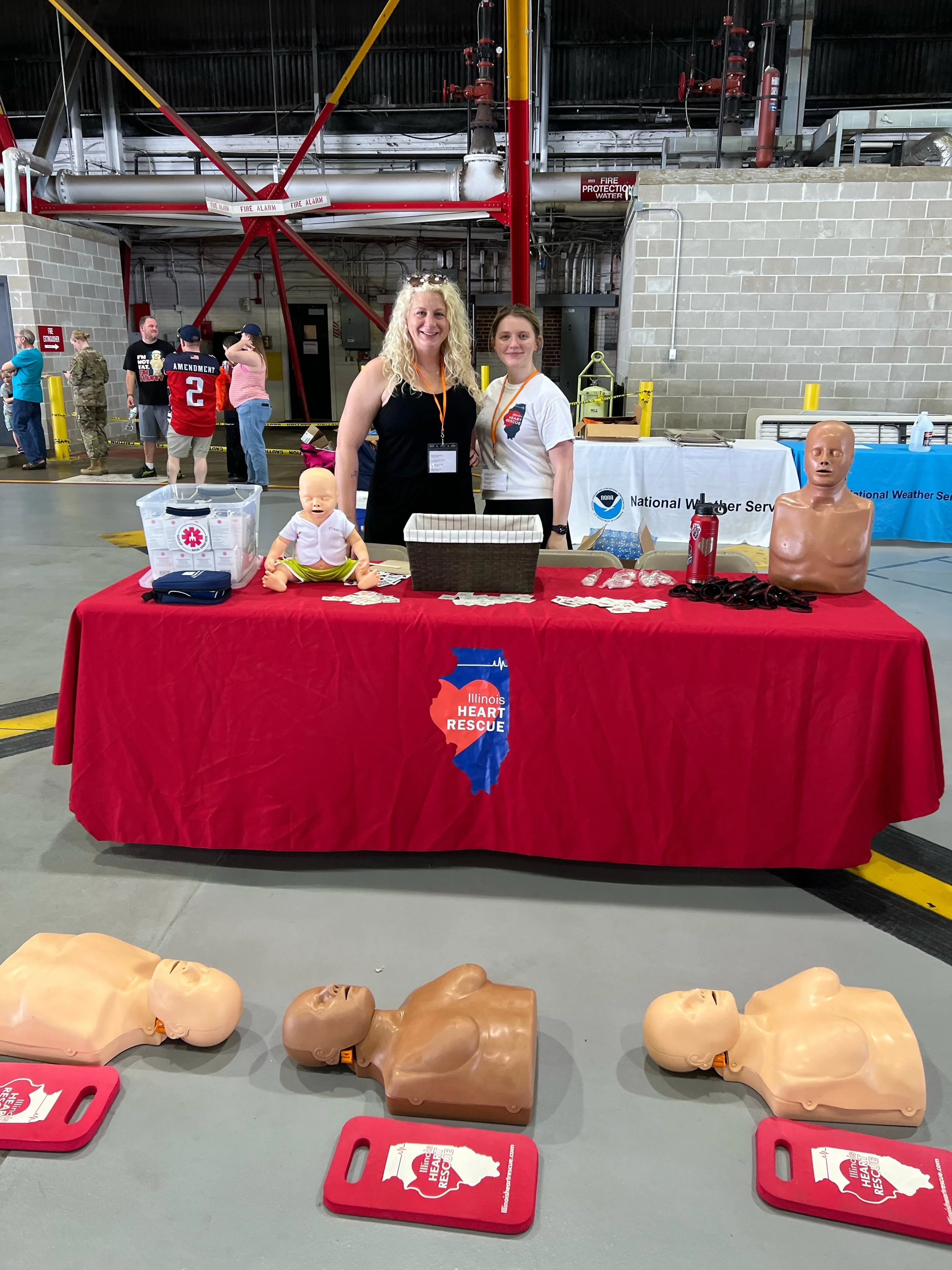 Table with Illinois Heart Rescue promotional materials, including child mannequins, AED trainers, and informational pamphlets, in a large industrial space with people in the background.