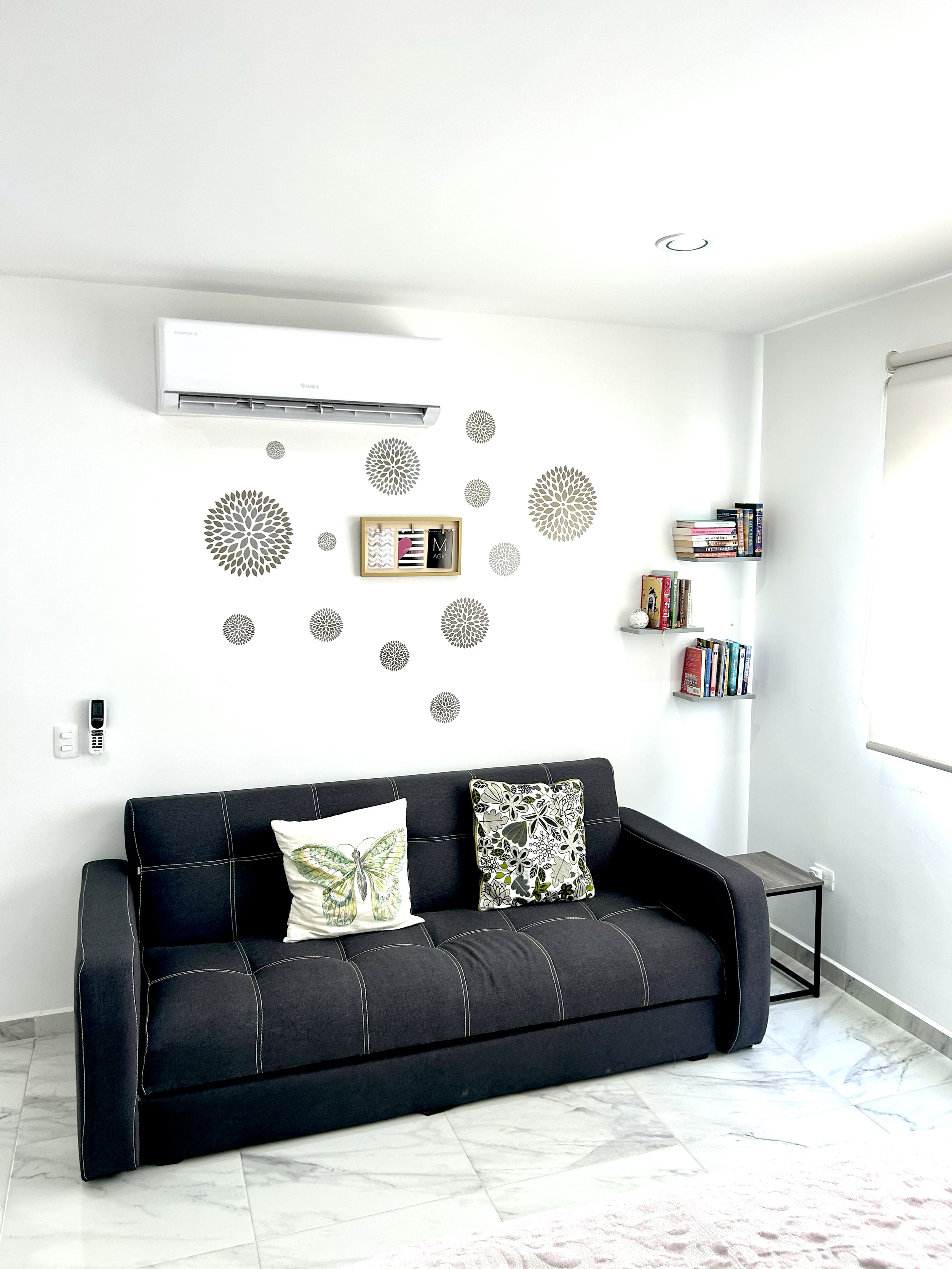 Living room with white walls, black sofa with floral and butterfly pillows, wall decor with gray floral patterns, wall-mounted air conditioner, and corner shelves with books.