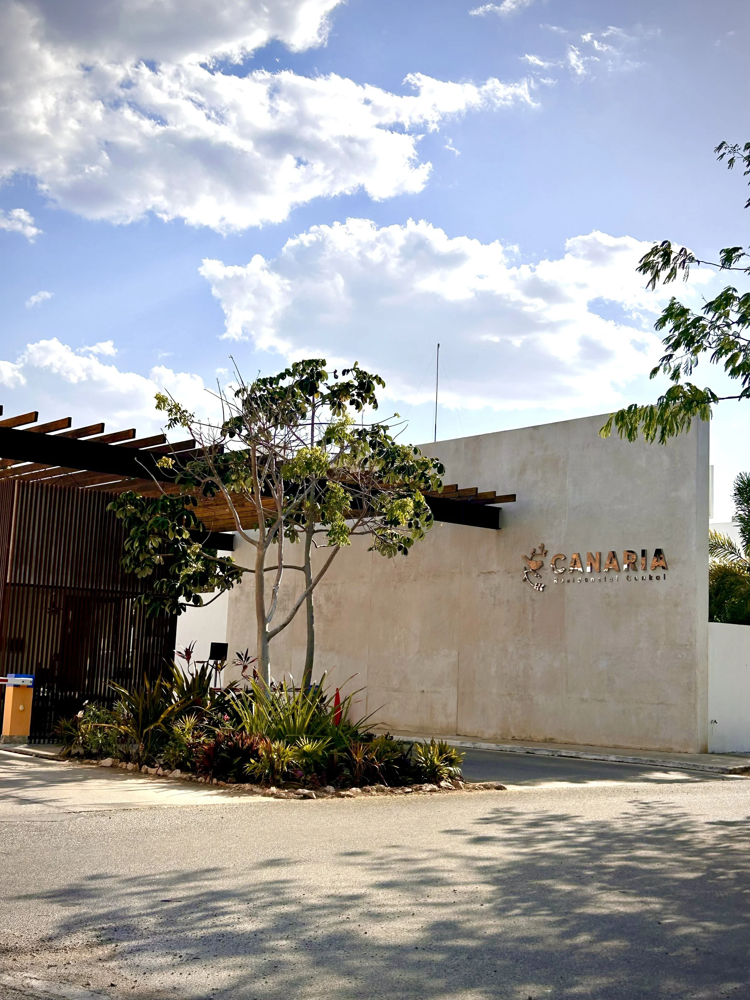 Entrance to a residential complex named Canaria, with a modern white wall, decorative metal sign, landscaped area with a tree and plants, and a clear blue sky with clouds.