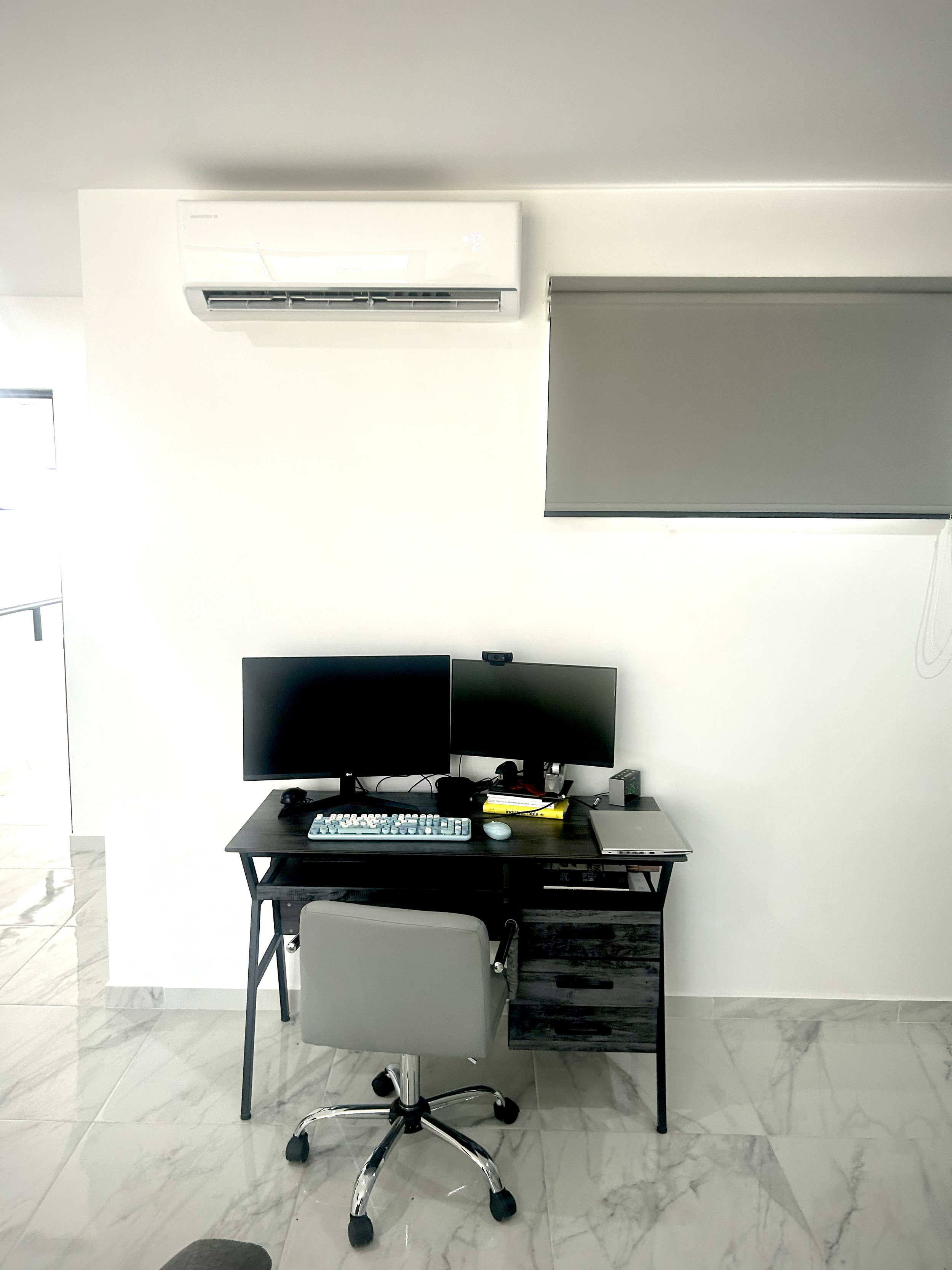 A home office with a black desk holding two computer monitors, a keyboard, and a mouse. There is a gray swivel chair in front of the desk. An air conditioning unit is mounted above, and a window with a gray shade is to the right.