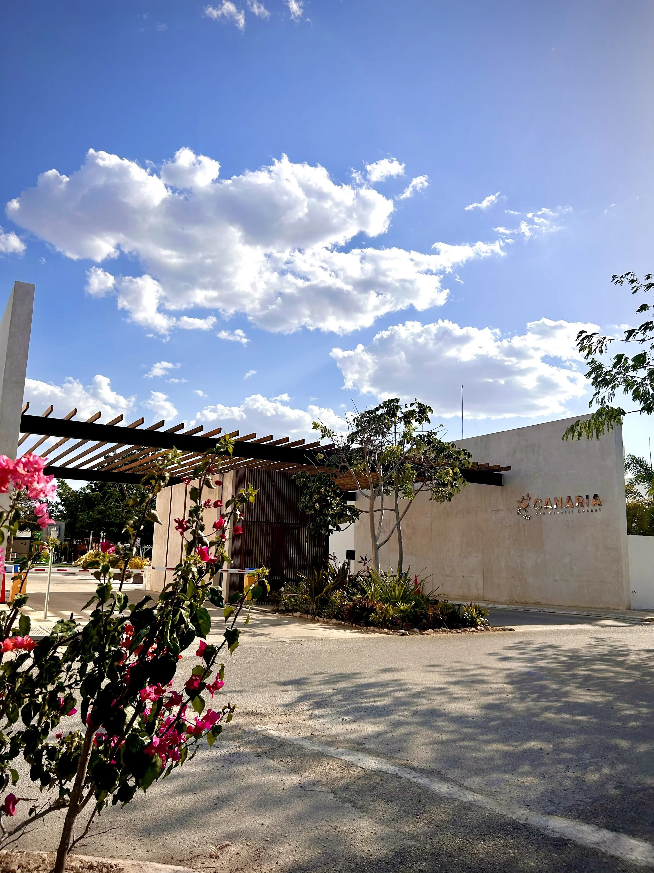 Entrance to Canara, a residential community or development, with a modern building, trees, and flowering bushes under a partly cloudy sky.