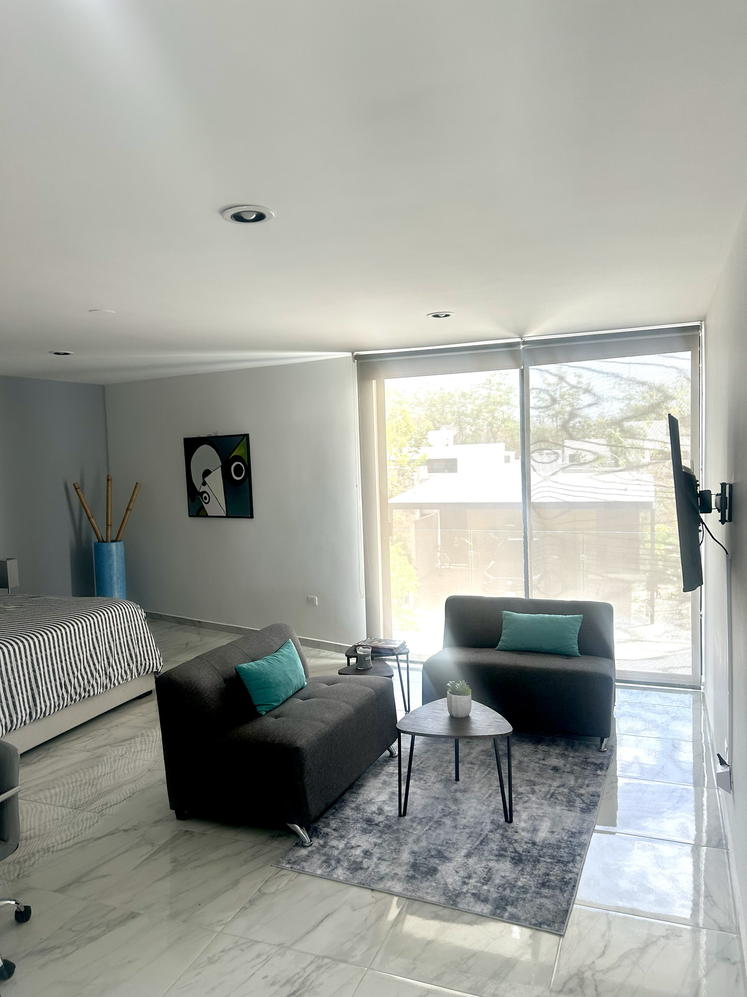 Living room with gray couch, black armchair, small coffee table, gray area rug, large windows with blinds, wall-mounted TV, decorative pillows, and an art piece on the wall.
