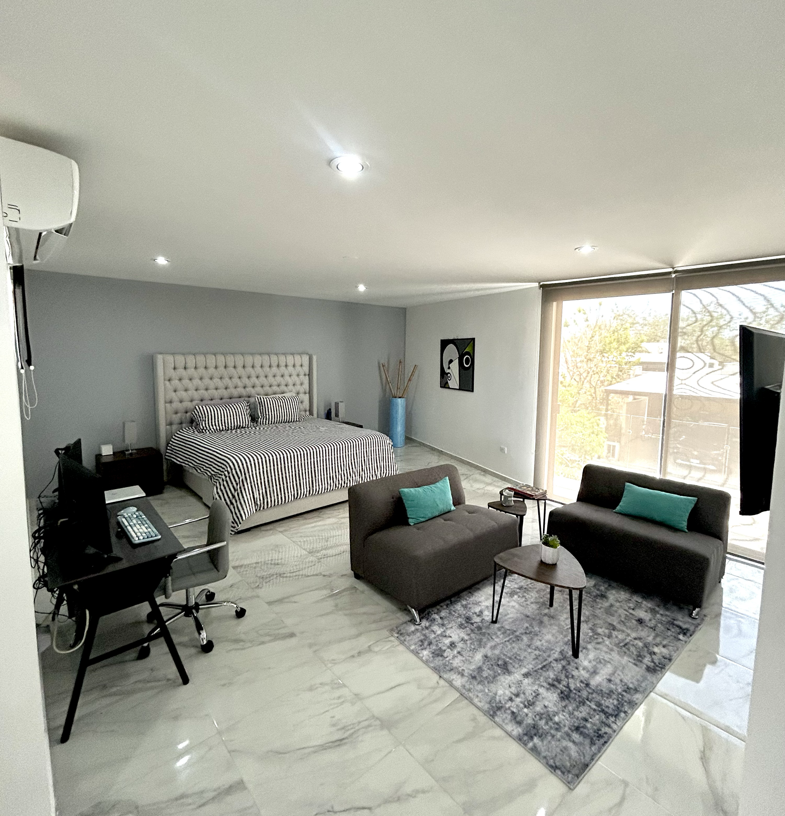 Bright modern studio apartment with a bed, two sofas, a small table, a workspace with a computer, and large sliding glass doors leading to a balcony.