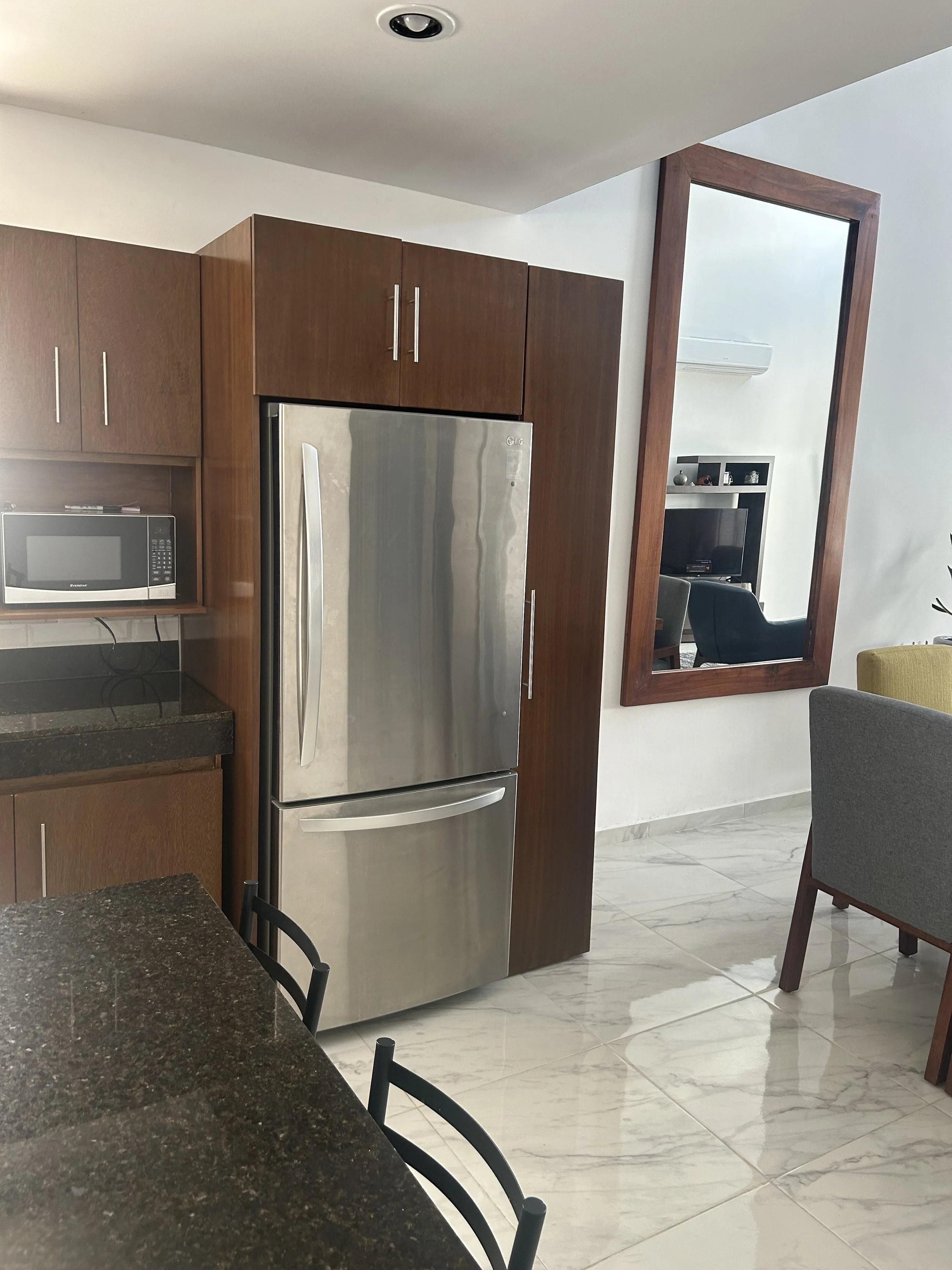A kitchen area with a stainless steel refrigerator, wooden cabinets, a microwave, and a black countertop. There is a large mirror on the wall reflecting a living room with television and chairs.