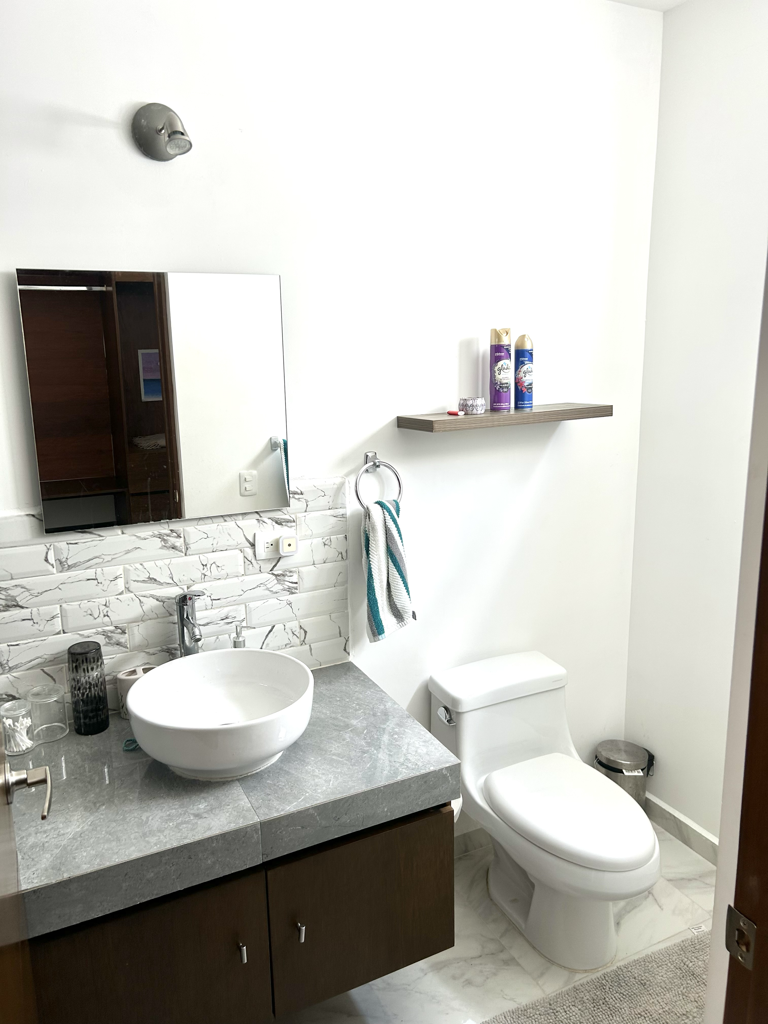 Bathroom with white walls, marble countertop with vessel sink, wall mirror, towel ring with towel, toilet, wall shelf with toiletries, small trash can, and light fixtures.