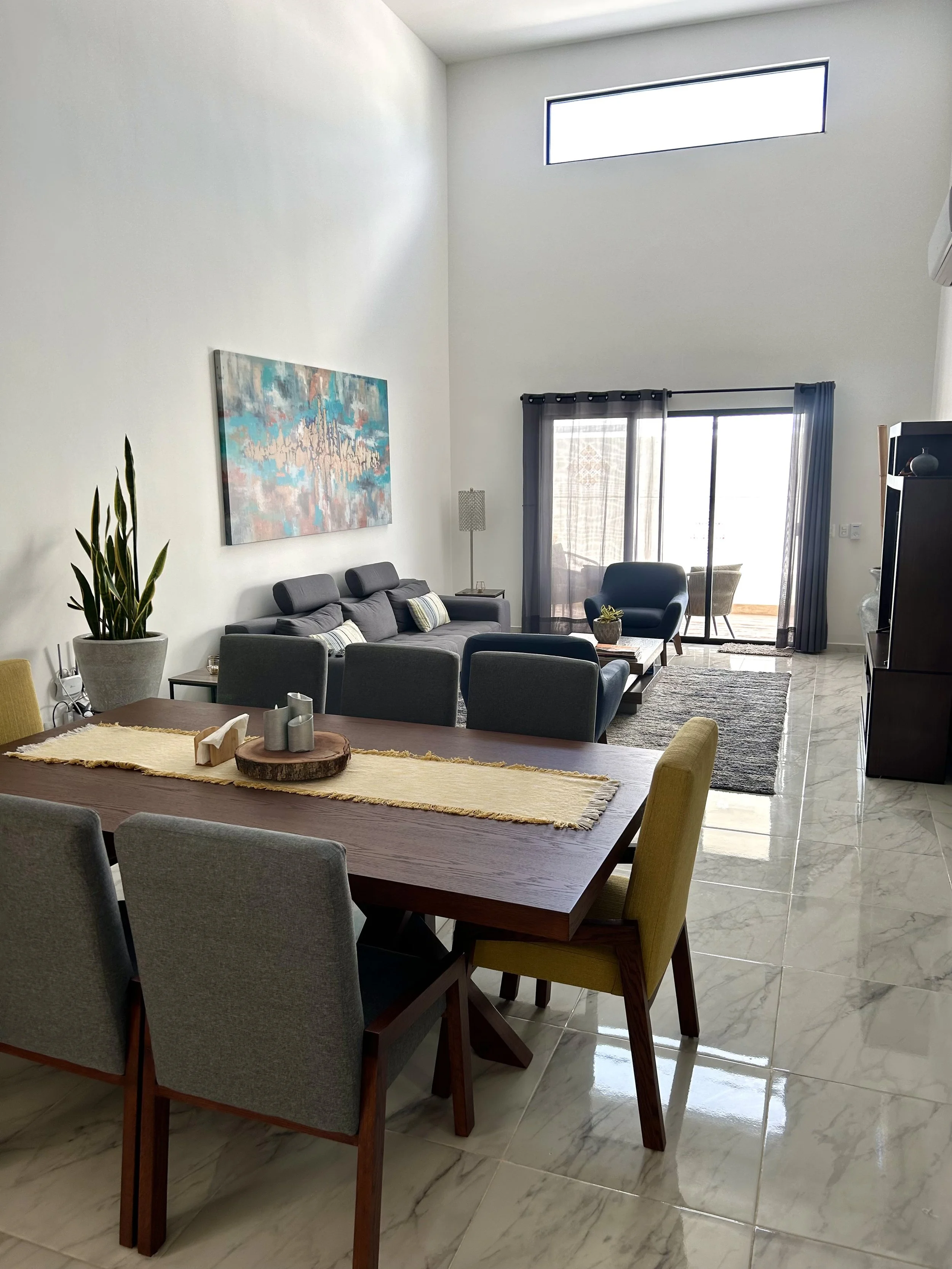 Living room with dining table, chairs, sofa, armchair, coffee table, sliding glass door with curtains, large windows, and tiled flooring.