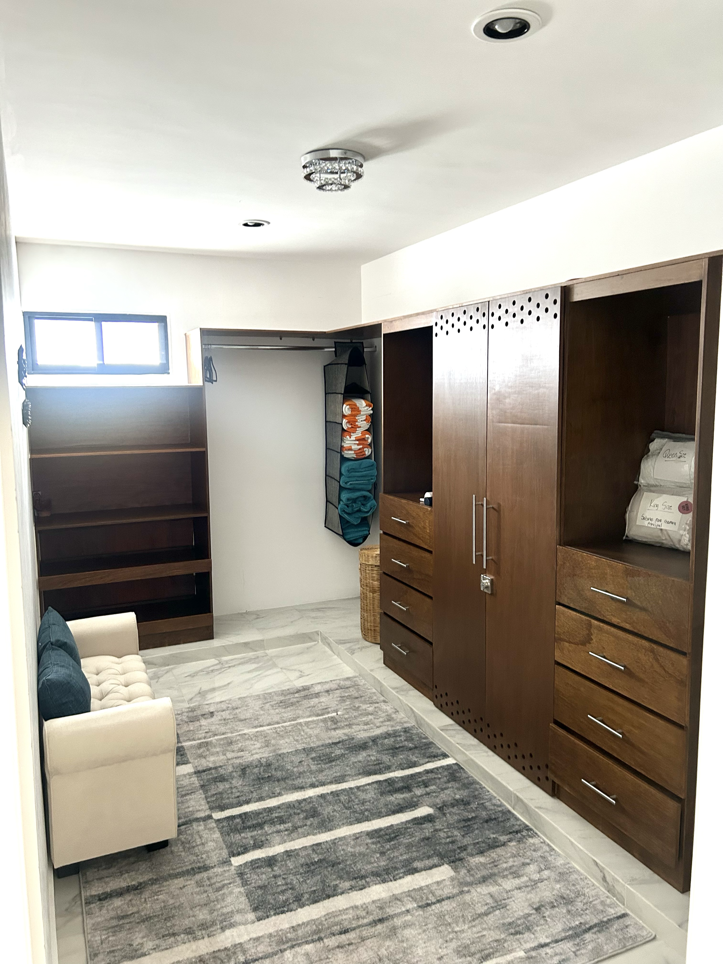 A room with white walls, a window near the ceiling, a beige armchair with a dark pillow, a patterned area rug, and wooden storage units with drawers and open shelves.