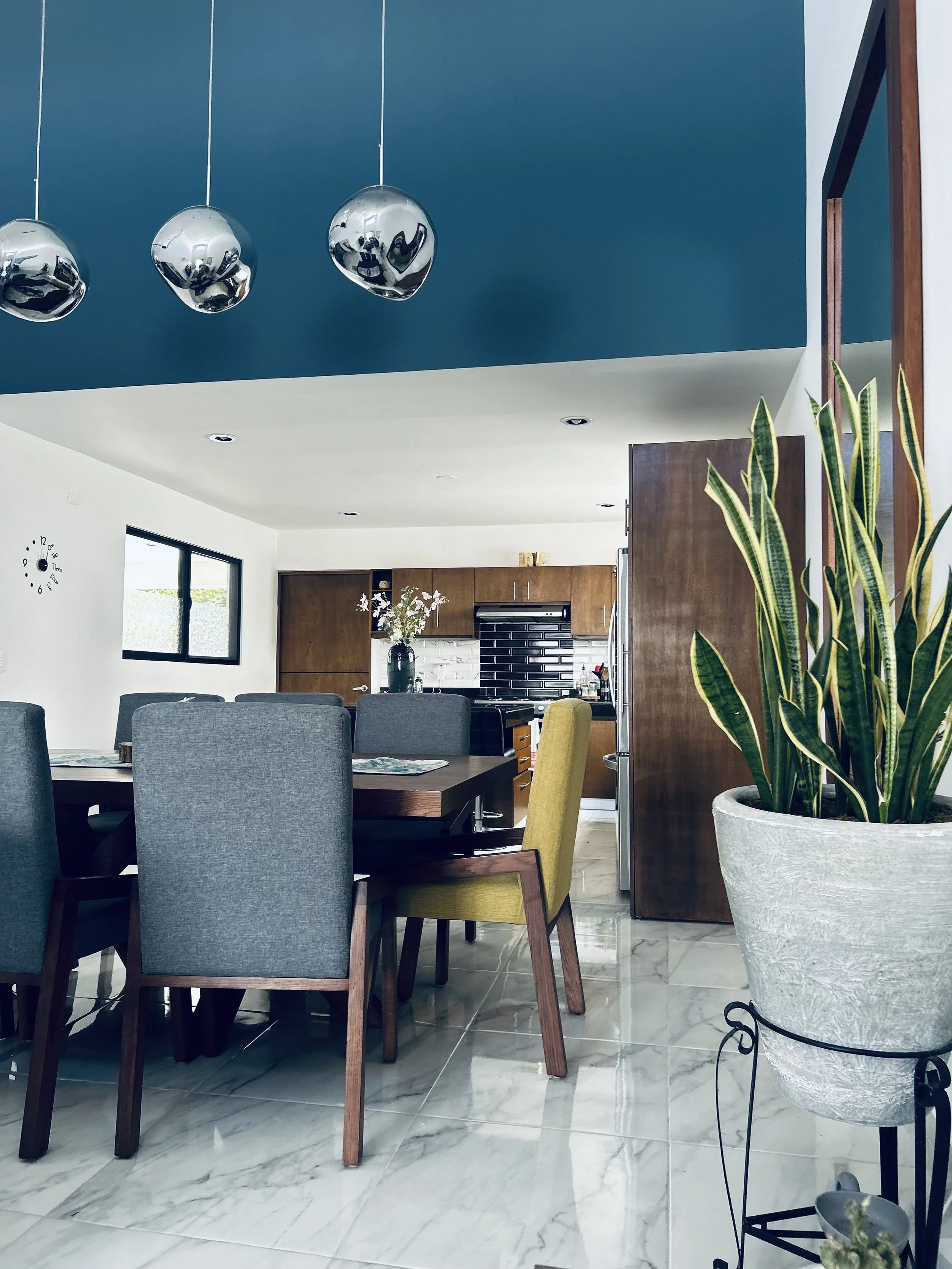 Modern dining area with a wooden dining table, upholstered chairs in gray and yellow, large potted plant, and a view of the kitchen with wooden cabinets and black backsplash. Three chrome pendant lights hang from the ceiling.